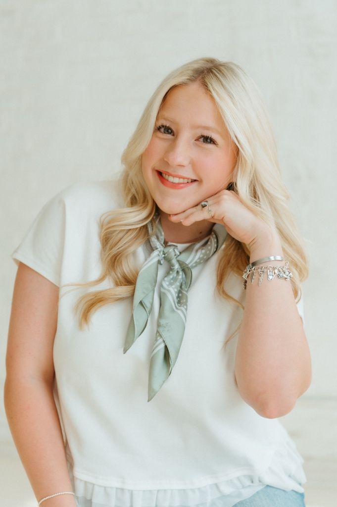 Smiling blonde woman in white top and sage green neck scarf poses with hand on chin in bright studio setting.