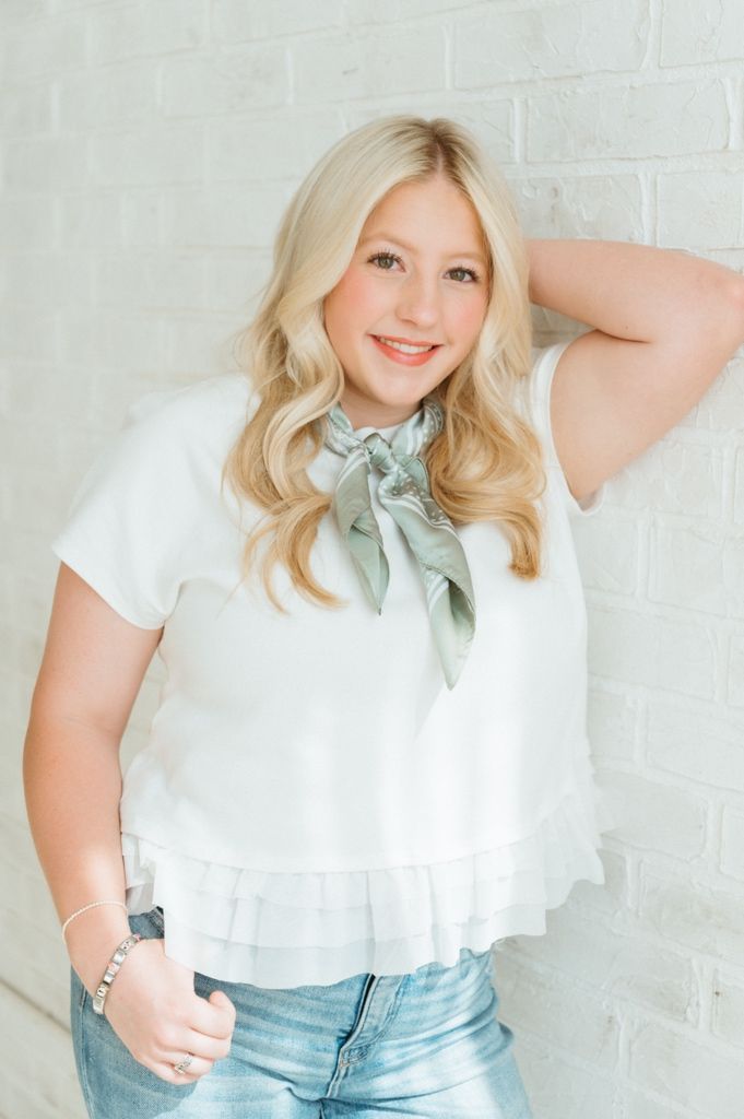 Blonde woman in white top with green neck scarf posing confidently against a white brick wall.