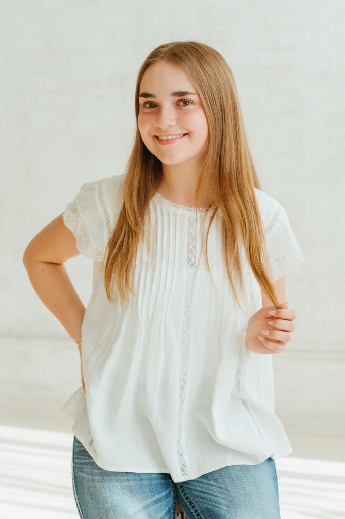 Smiling teenage girl with long blonde hair wearing a white lace blouse and jeans, posing against a light gray wall.