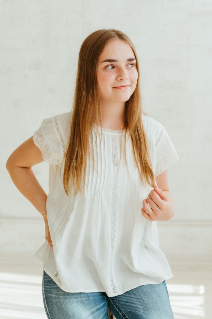 Young woman with long hair wearing a white lace blouse and jeans posing indoors.