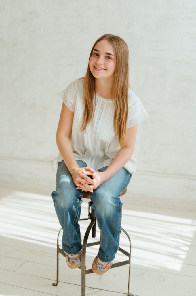 Smiling teenage girl in white blouse and jeans sitting on a stool in bright studio.