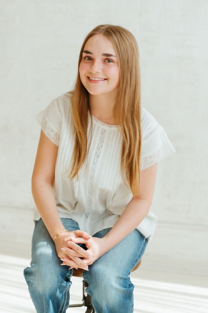 Smiling teenage girl with long blonde hair sitting on a stool, wearing a white blouse and blue jeans.