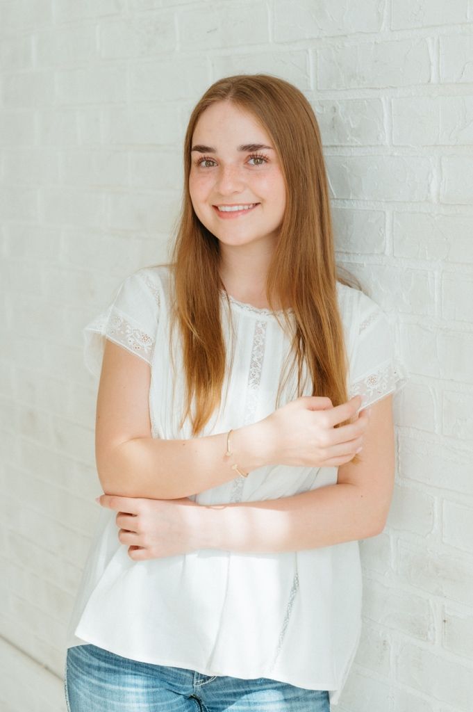Smiling young woman with long brown hair in white blouse and jeans posing against white brick wall.