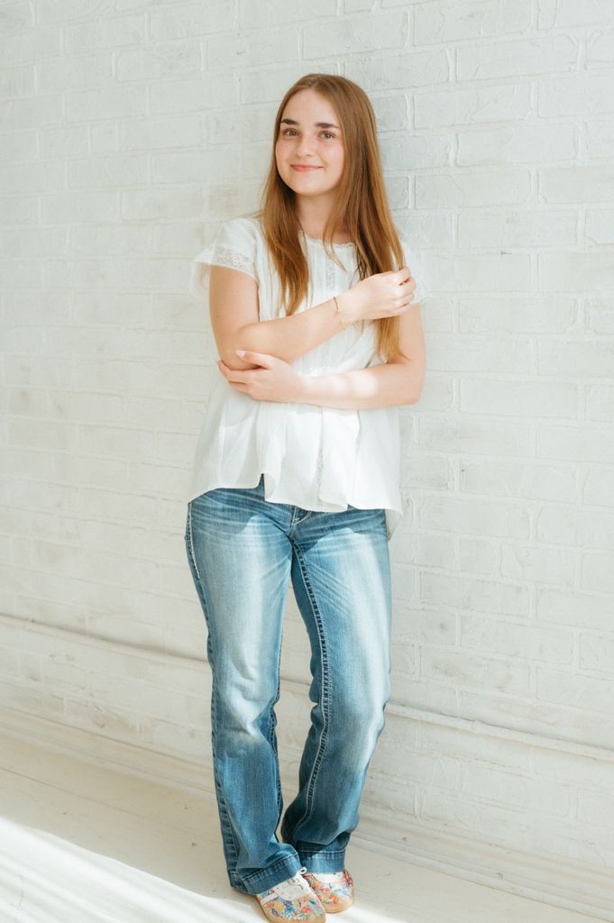 Young woman with long brown hair in white top and blue jeans leaning against white brick wall.