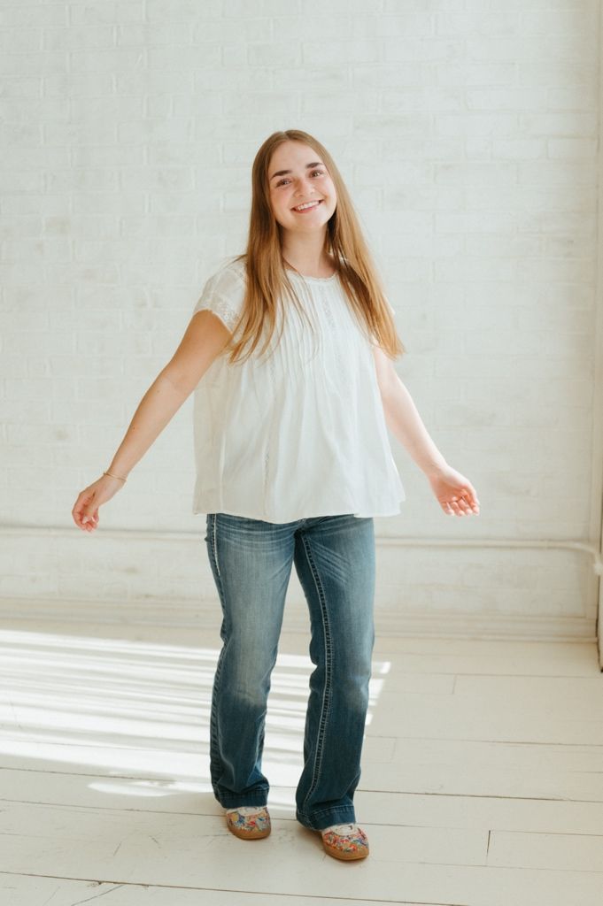 Smiling teenage girl with long blonde hair wearing a white blouse and blue jeans standing in a bright white studio.
