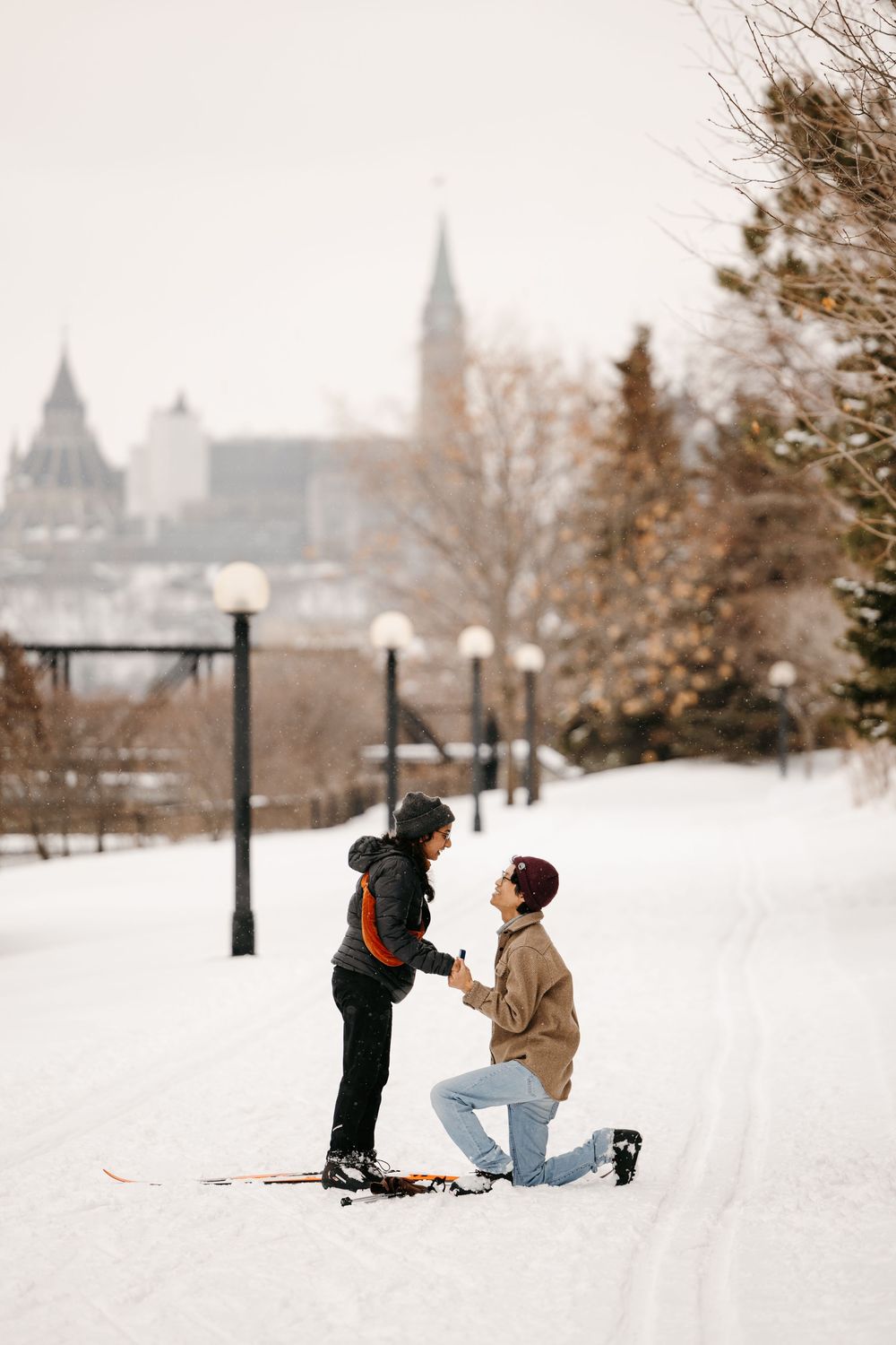 Couple sharing a romantic marriage proposal on a snowy path in winter park with historic Ottawa skyline.