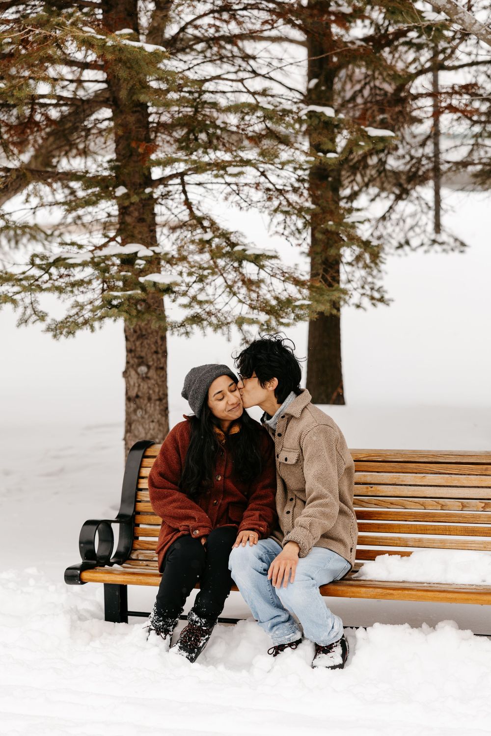 Romantic couple sharing a kiss while sitting on a wooden bench in a snowy winter park.