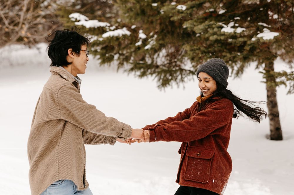 Happy couple holding hands and spinning together in a snowy winter park with evergreen trees.
