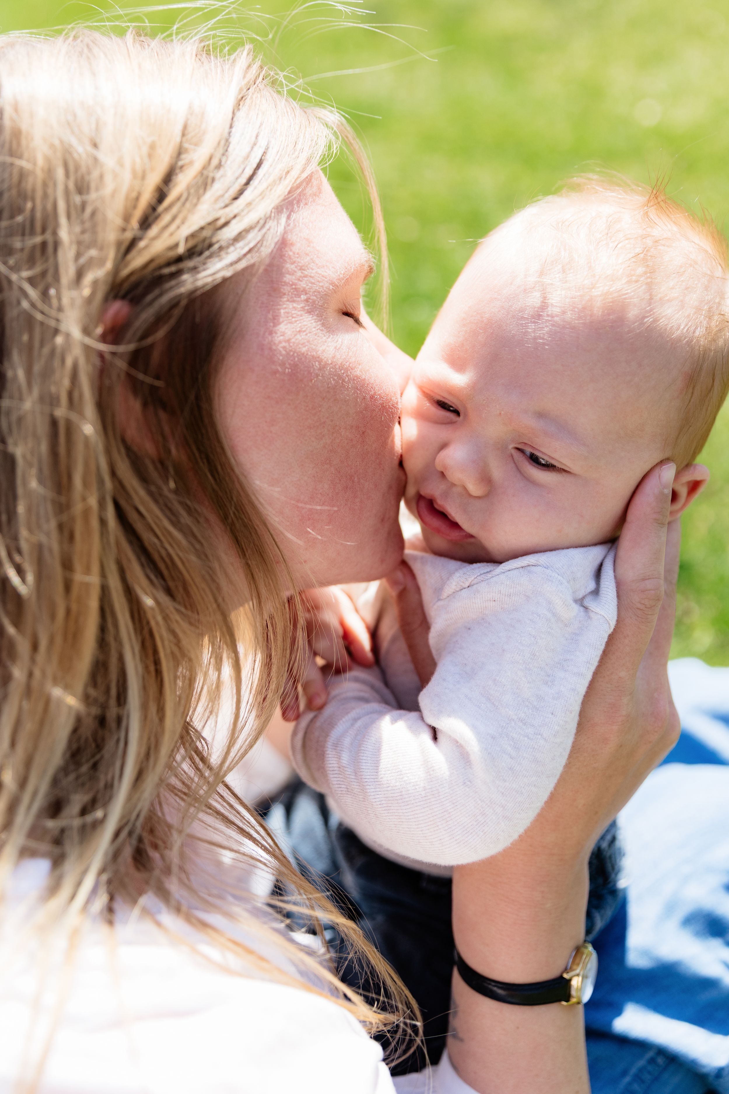 A mother tenderly kisses her smiling baby outdoors on a sunny day, holding the infant gently in her hands.