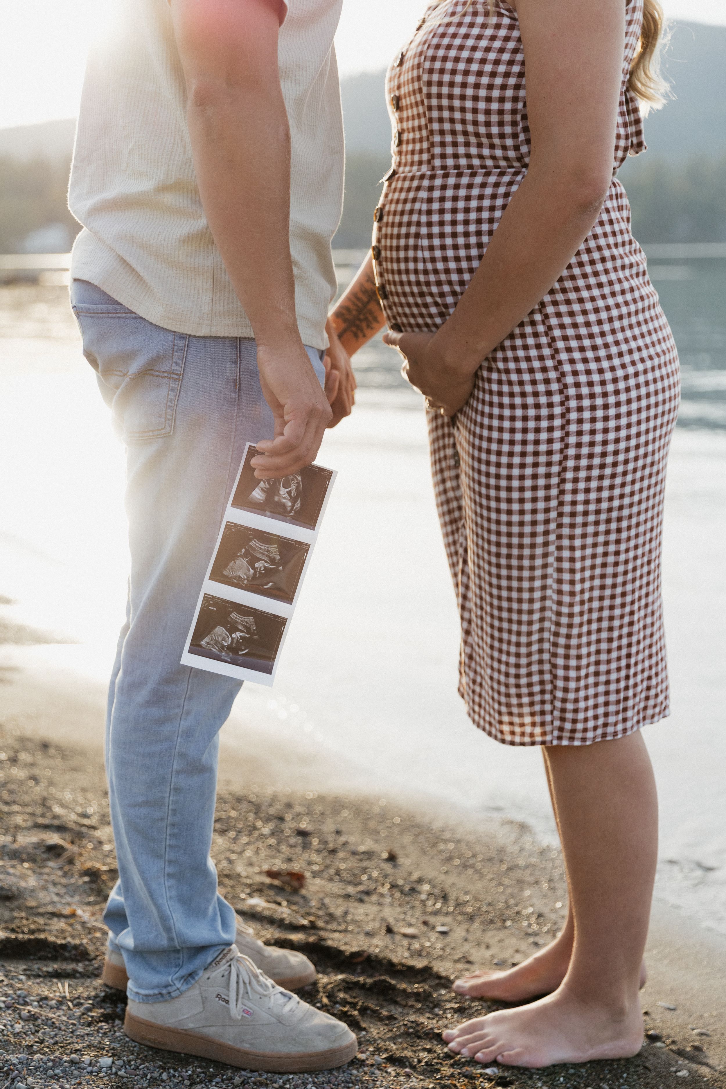Pregnant couple standing on beach holding ultrasound photos, woman in checkered dress, golden sunset light.