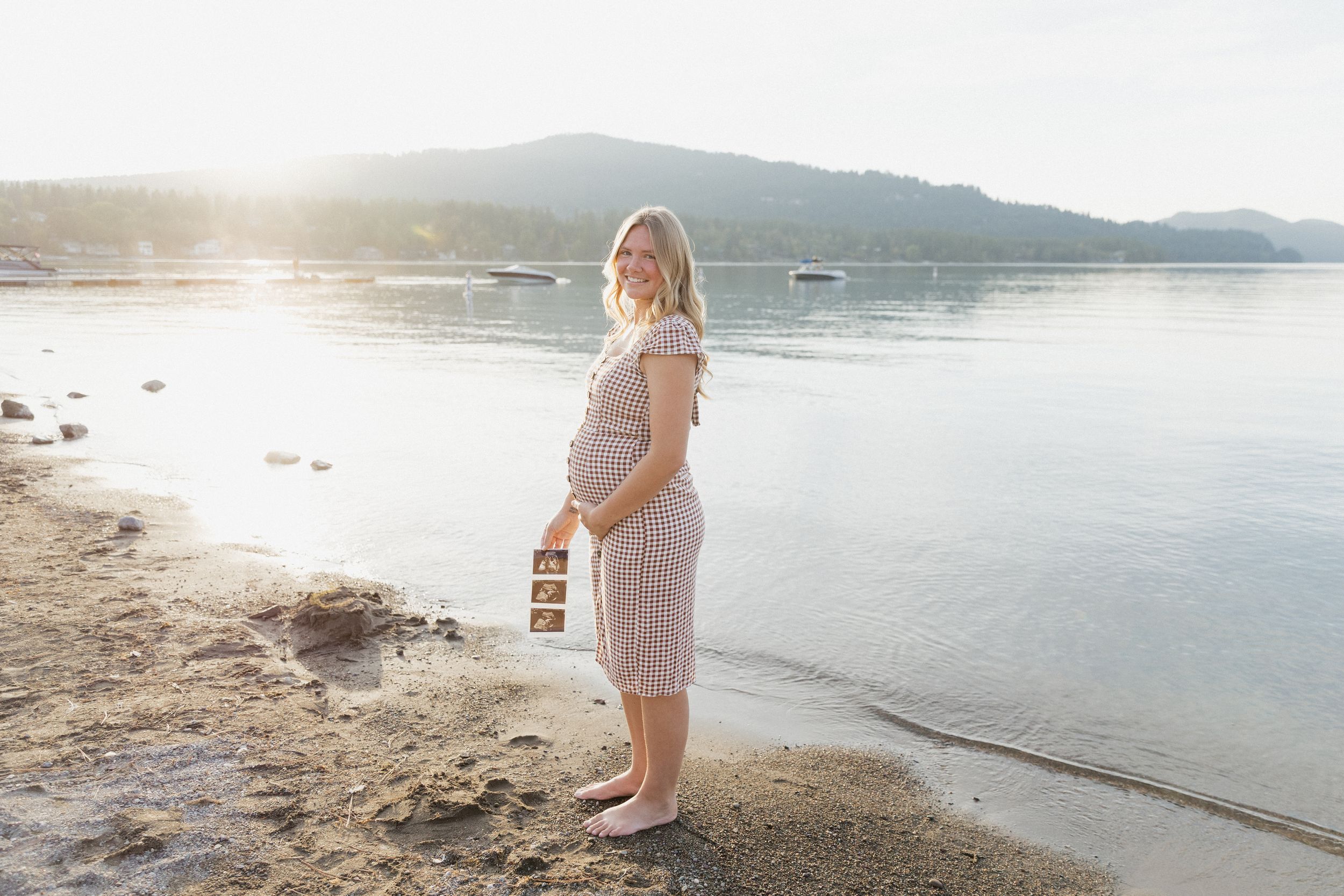 Pregnant woman in pink dress holding ultrasound photos on sunny beach shoreline with mountain backdrop.