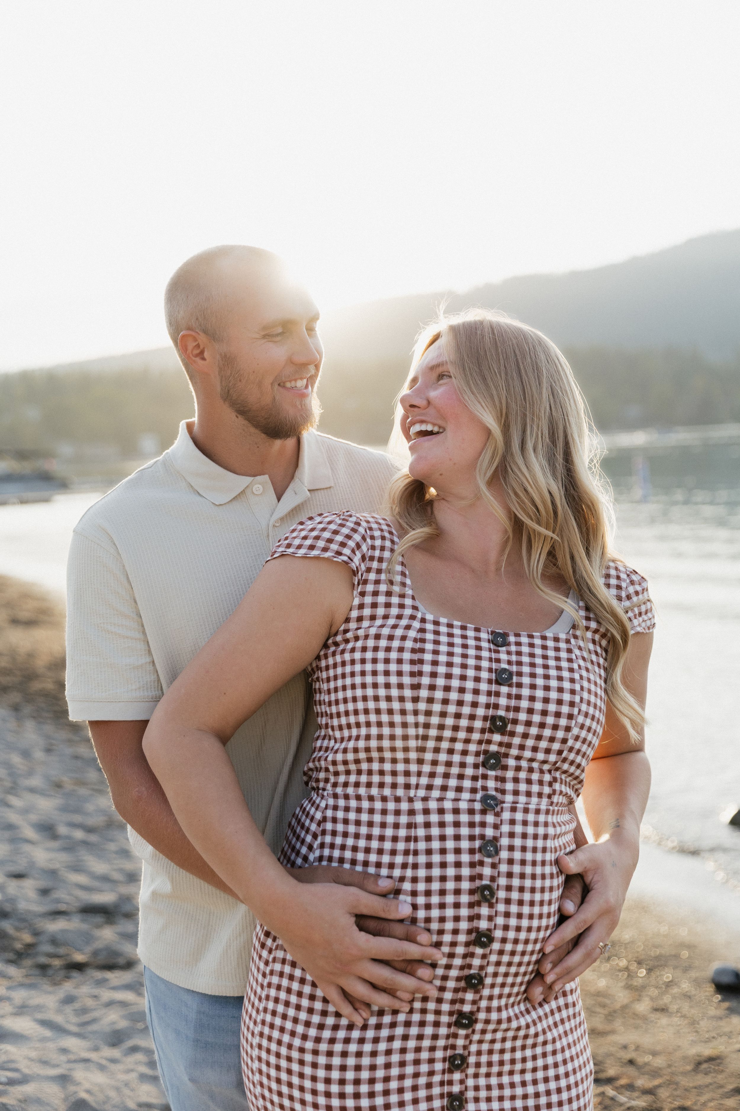 Smiling pregnant couple embracing on a sunny beach at golden hour, woman in plaid dress cradling baby bump.