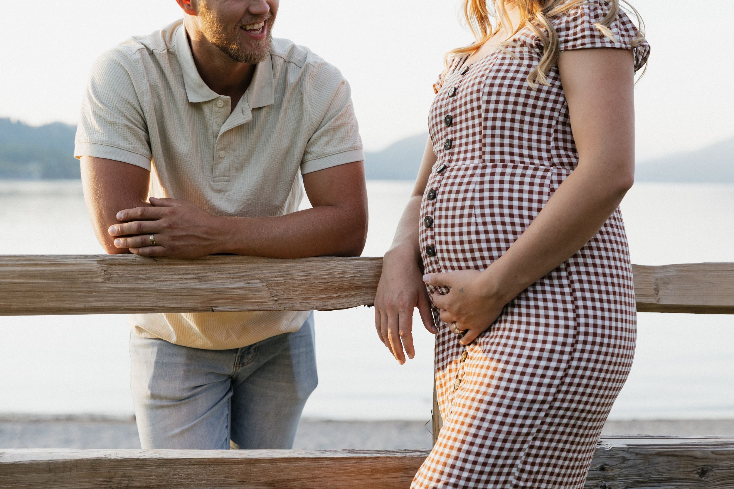 Pregnant woman in plaid dress with partner leaning on wooden railing by a lake during golden hour.