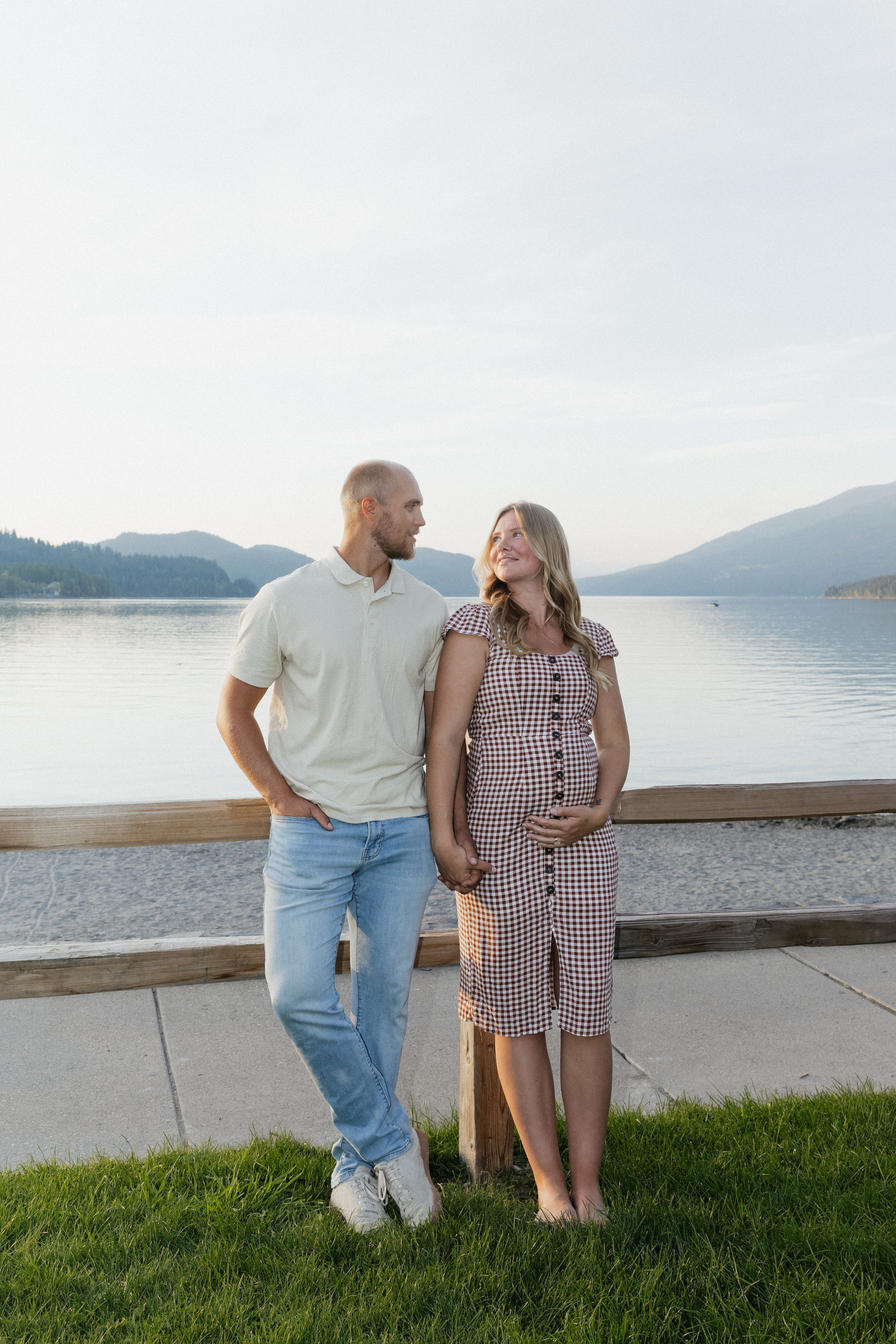 Expecting couple standing by a lakeside railing at dusk, woman in checkered dress holding her baby bump.
