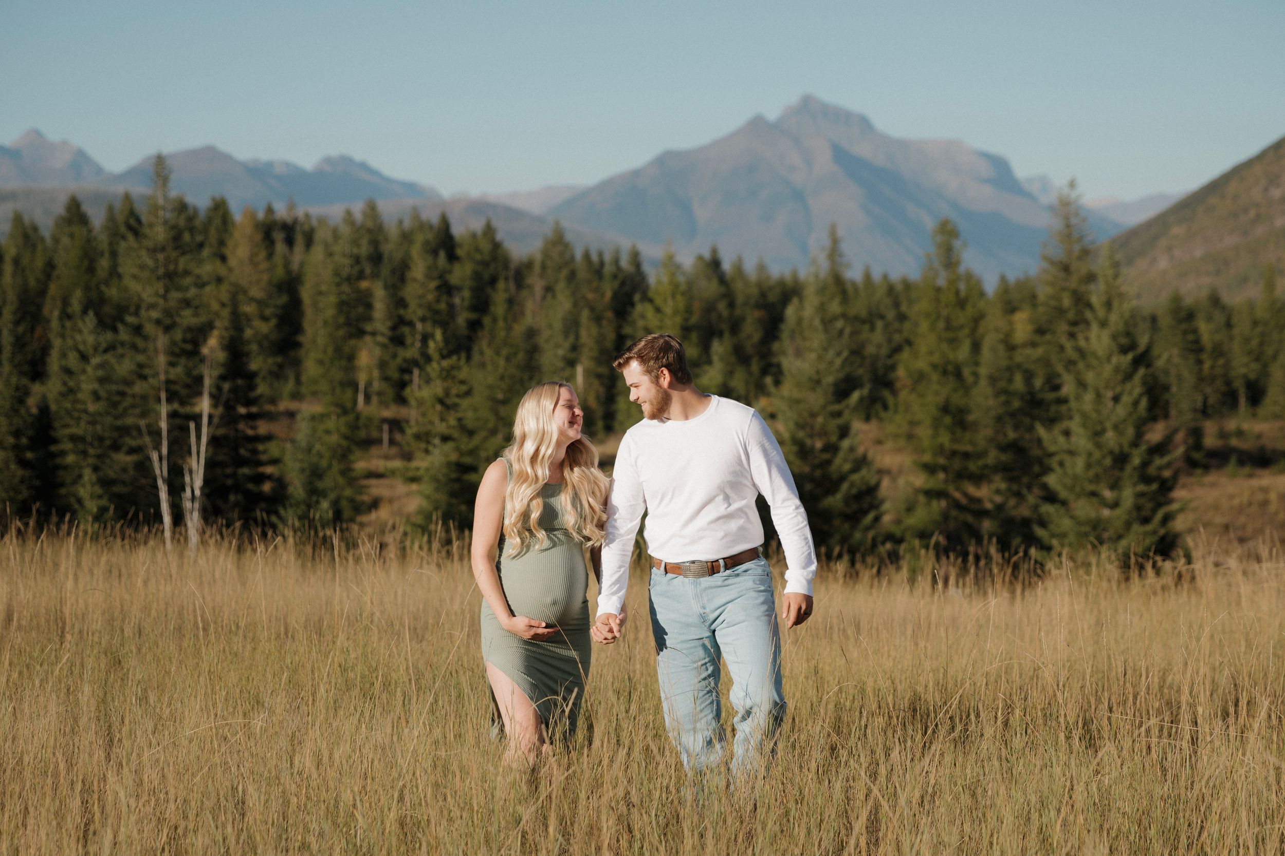 Pregnant couple holding hands in golden meadow with mountain backdrop and pine trees.