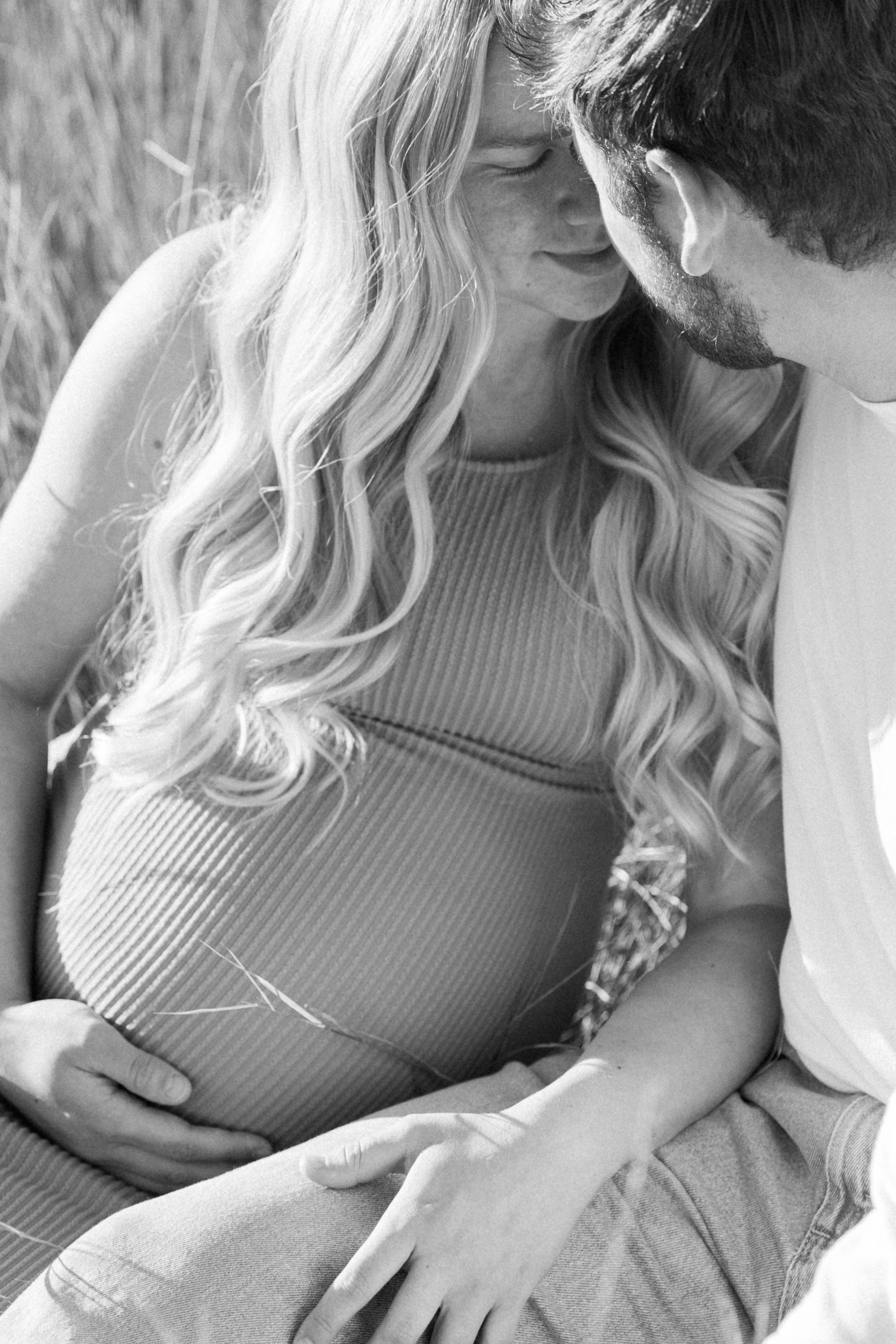 Black and white photo of a pregnant woman and her partner tenderly holding her baby bump outdoors.