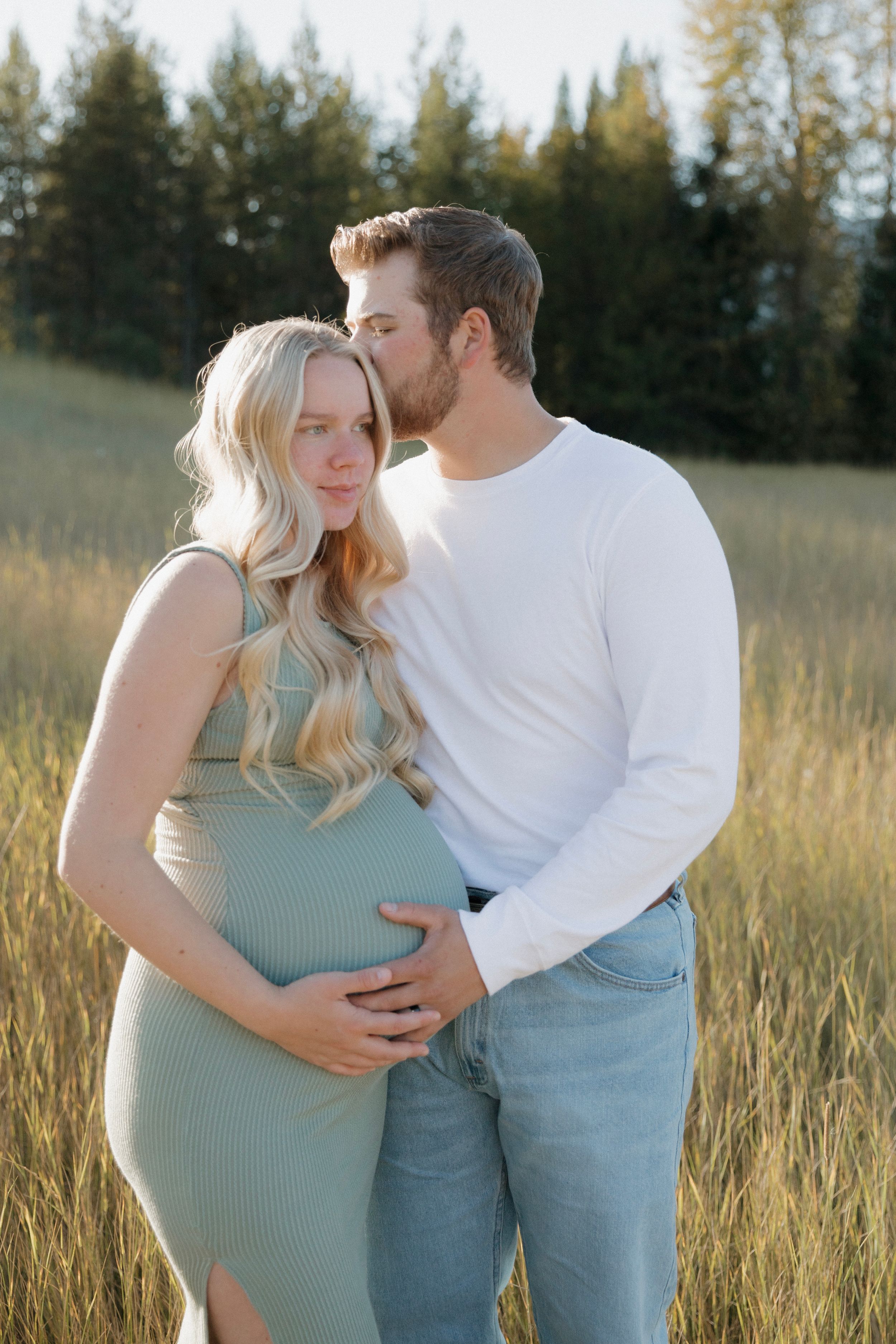 Pregnant couple embracing in golden field, man kissing woman's temple while cradling baby bump at sunset.