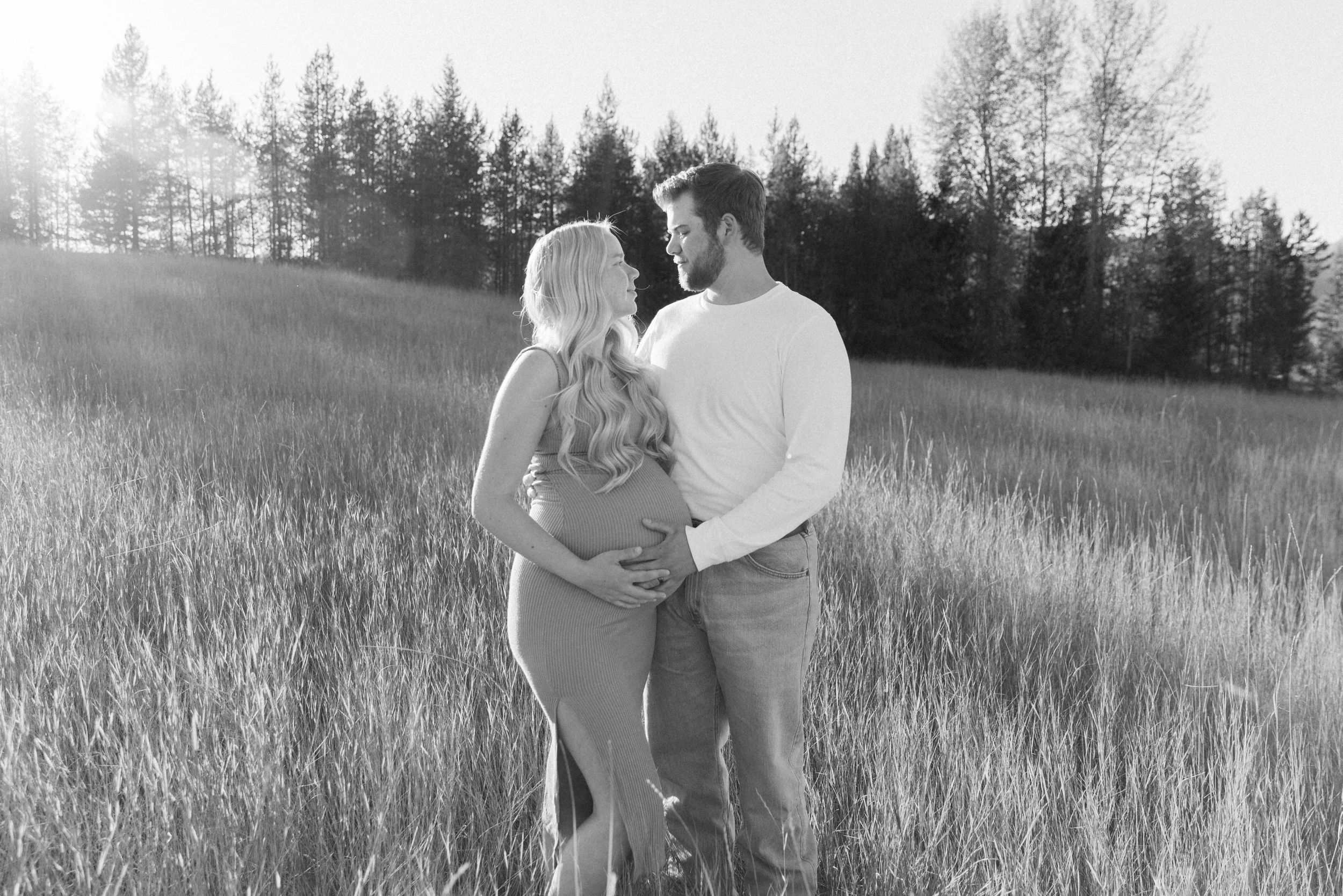 Black and white maternity photo of a couple embracing in a field with tall grass and trees in the background.