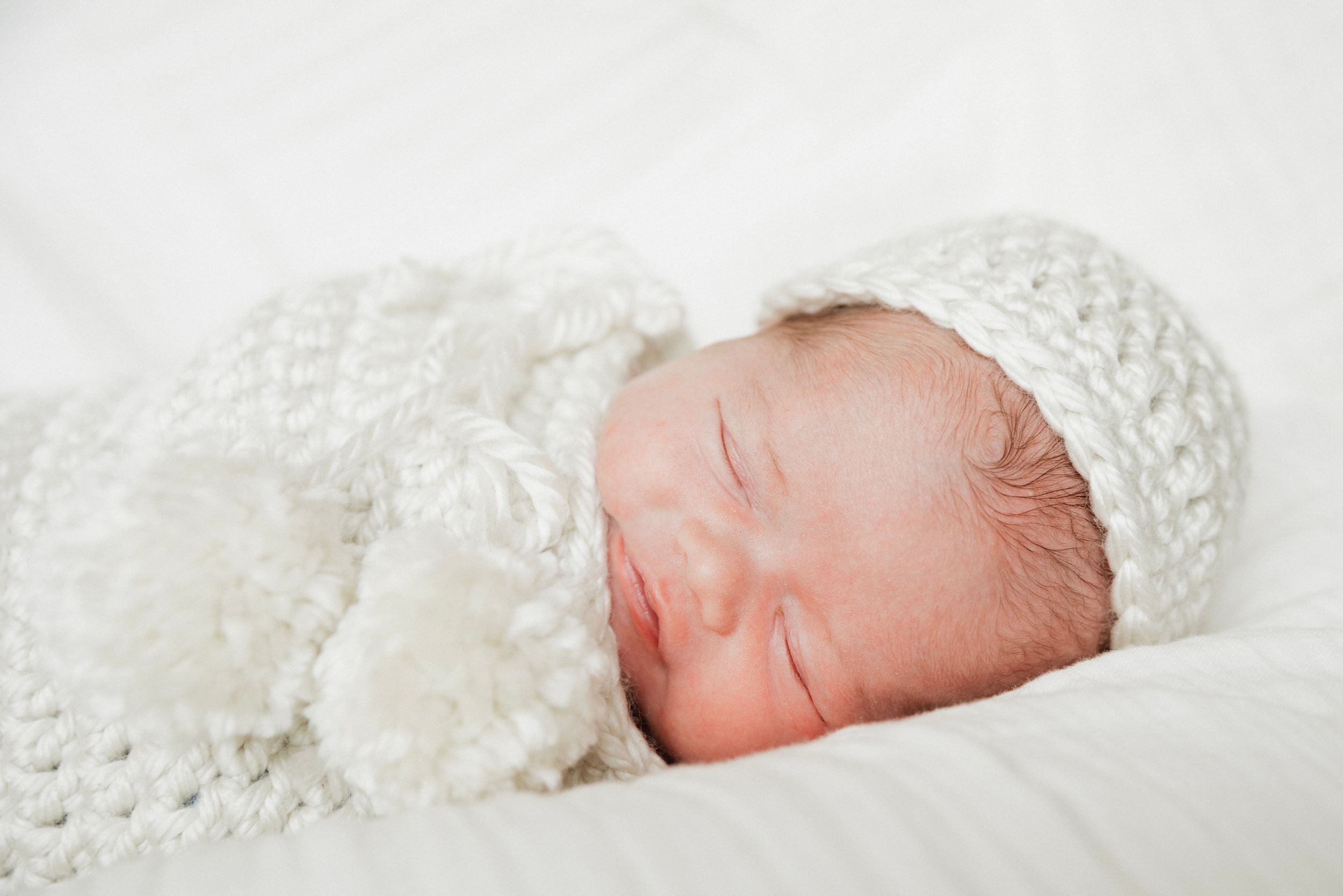 Sleeping newborn baby wrapped in a white knit blanket and matching crochet hat on a soft white background.
