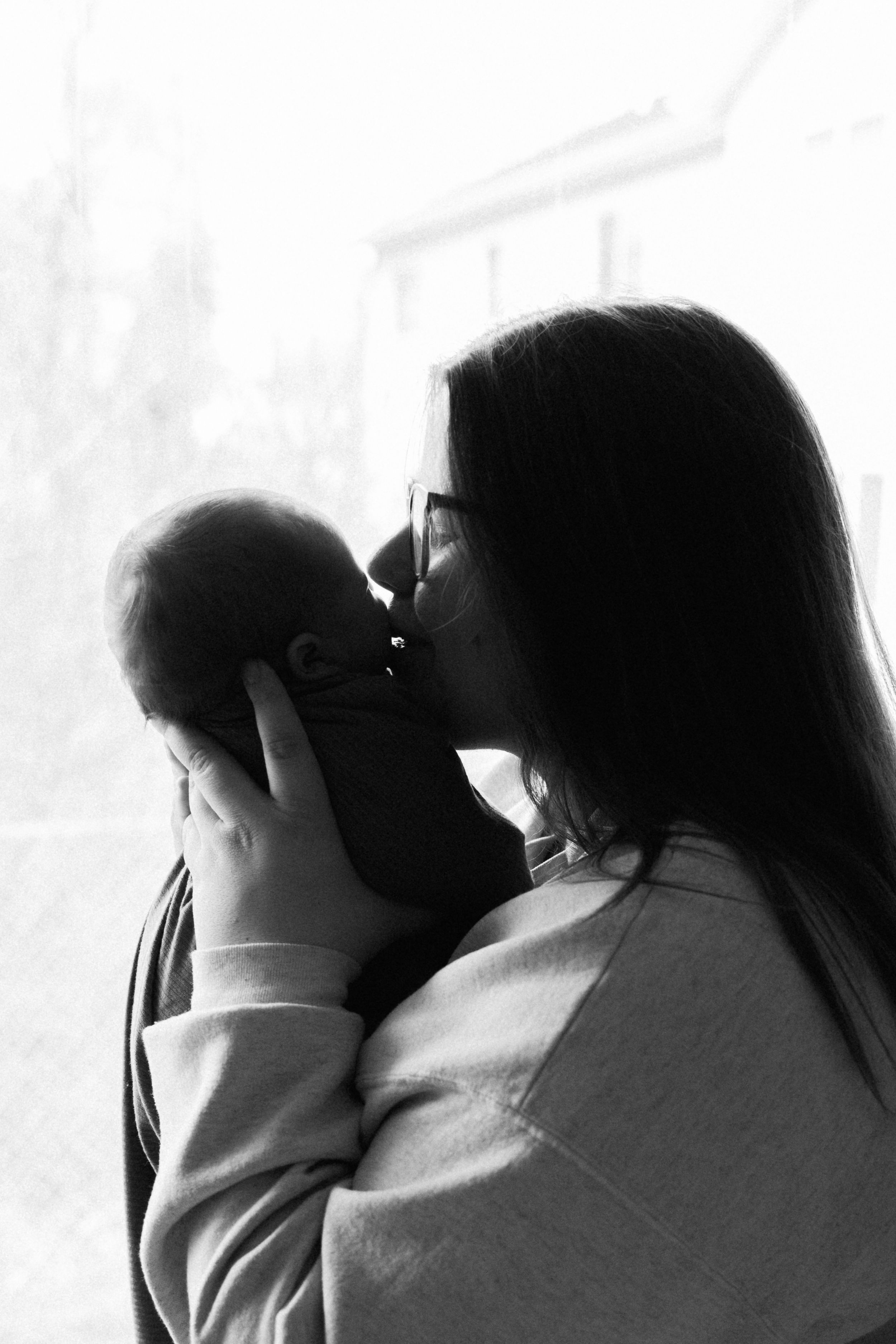 Black and white photo of a mother tenderly kissing her newborn baby while holding them near a bright window.