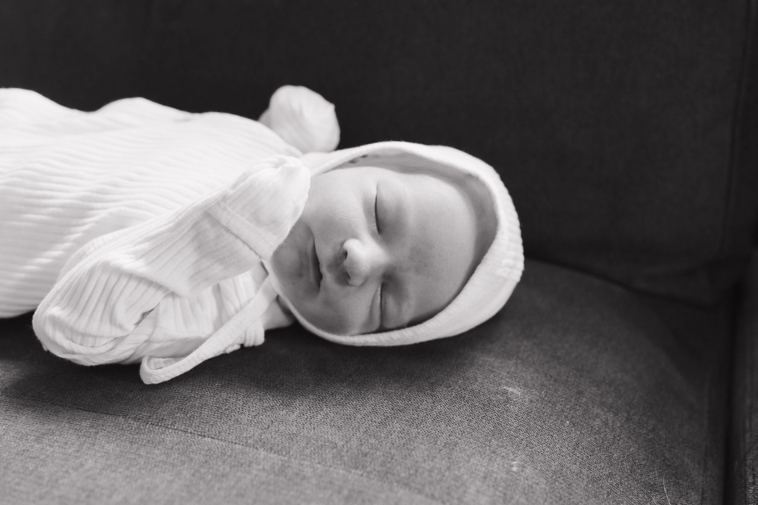 Black and white photo of a sleeping newborn baby wearing a white onesie and hat, resting on a gray surface.