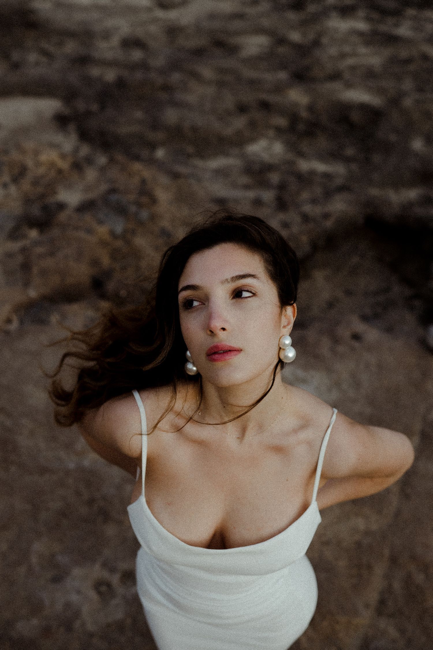Woman in white slip dress with pearl earrings and gold necklace posing on rocky terrain with wind-blown dark hair.