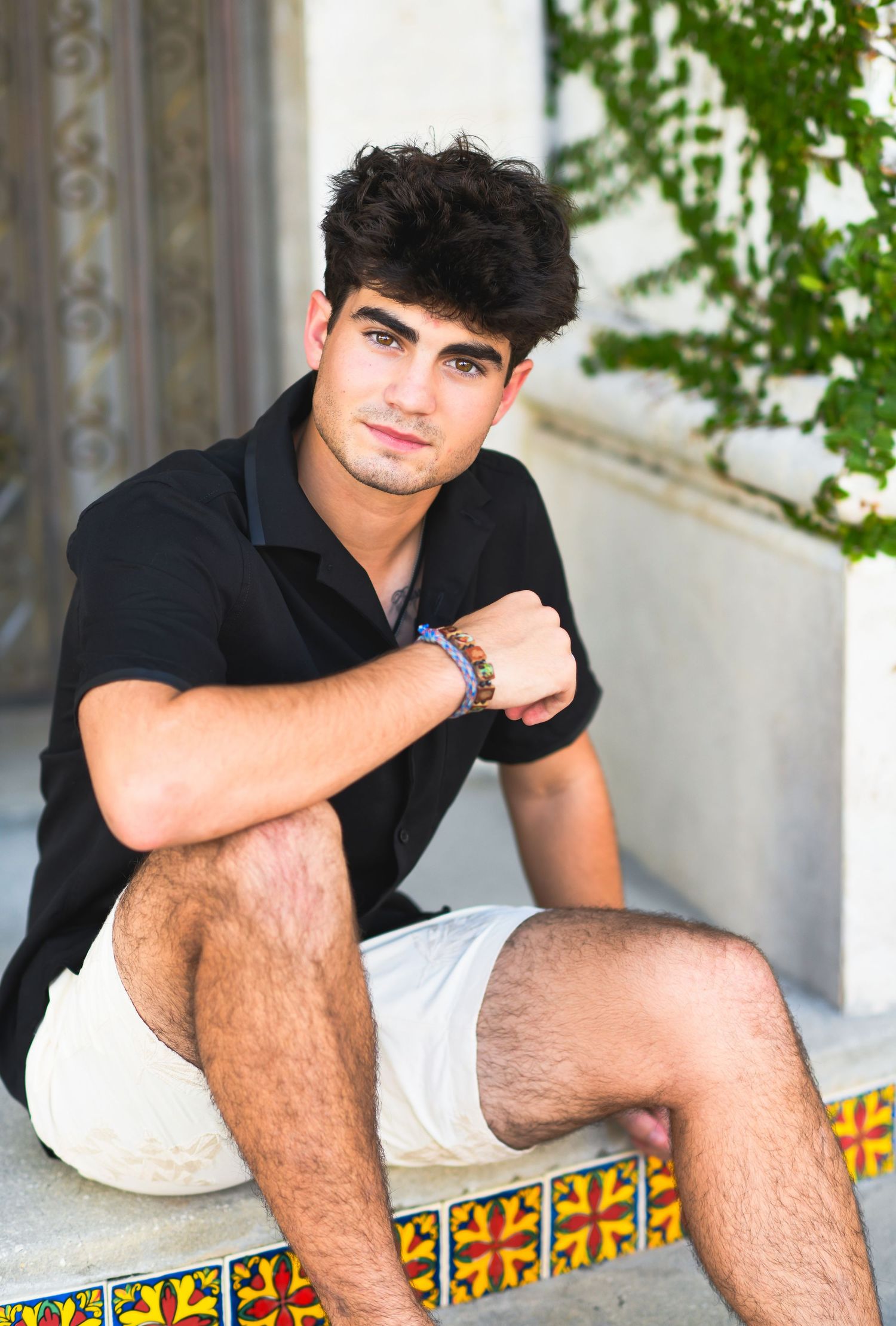 Young man with dark hair sitting on colorful tiled steps wearing black shirt and white shorts with beaded bracelets.
