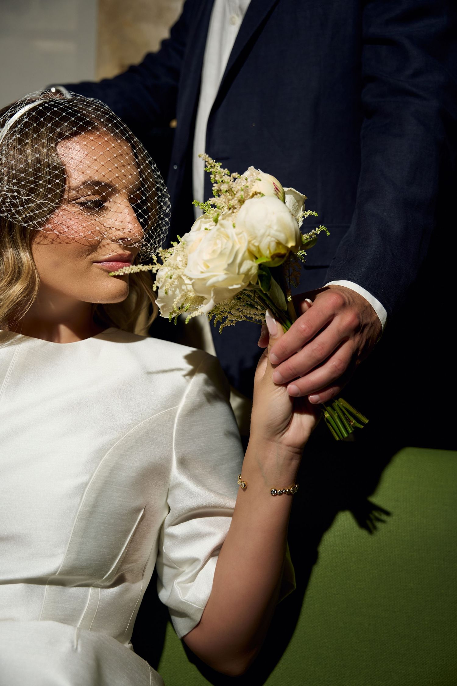 Bride in white dress and birdcage veil holds white rose bouquet with groom in navy suit.