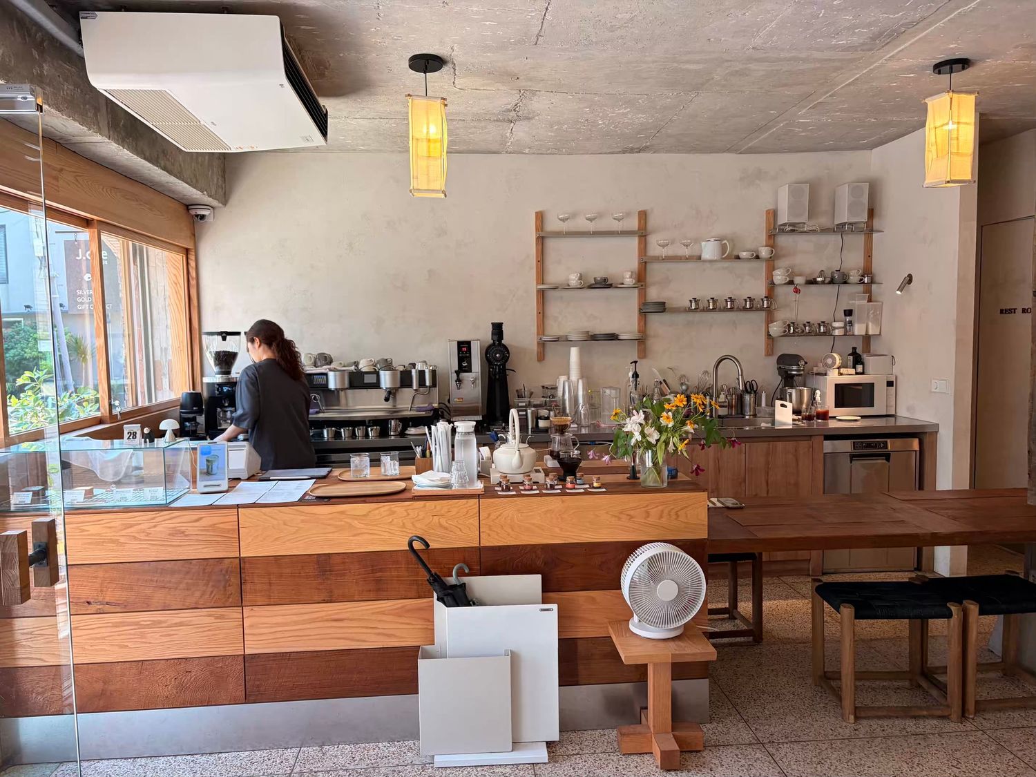 Modern minimalist cafe interior with wooden counter, barista, pendant lights, and open shelving with coffee equipment.