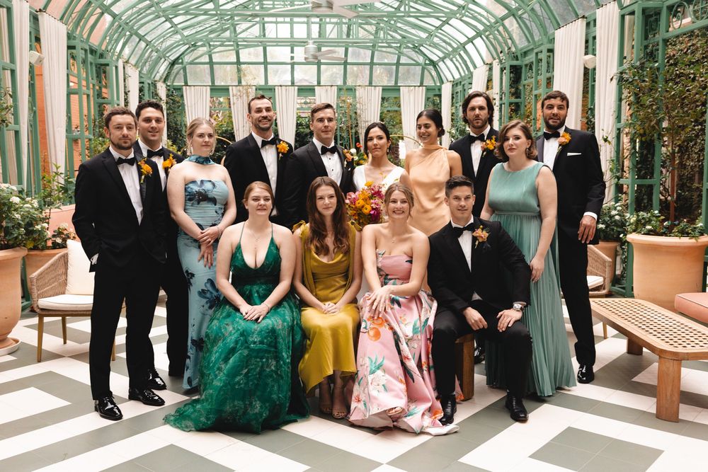 Elegant wedding party poses in a greenhouse venue, wearing formal attire in jewel tones including emerald, gold, and floral gowns.