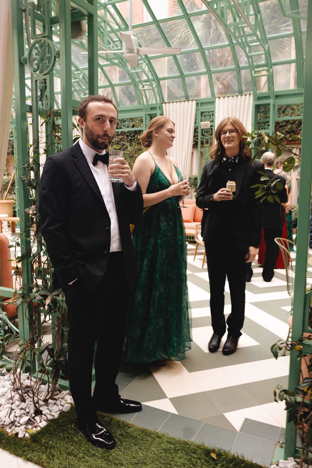 Elegant guests in formal attire socializing in a greenhouse venue with green iron framework and checkered floors.