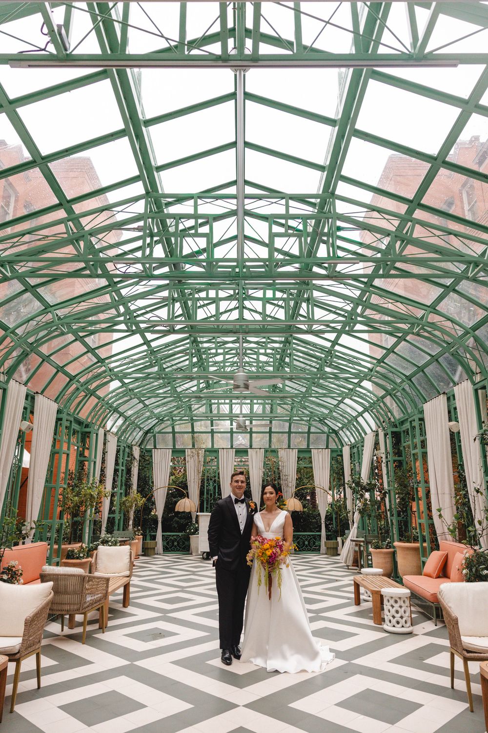 Bride and groom stand in elegant glass greenhouse conservatory with green steel arches and geometric tile floor.
