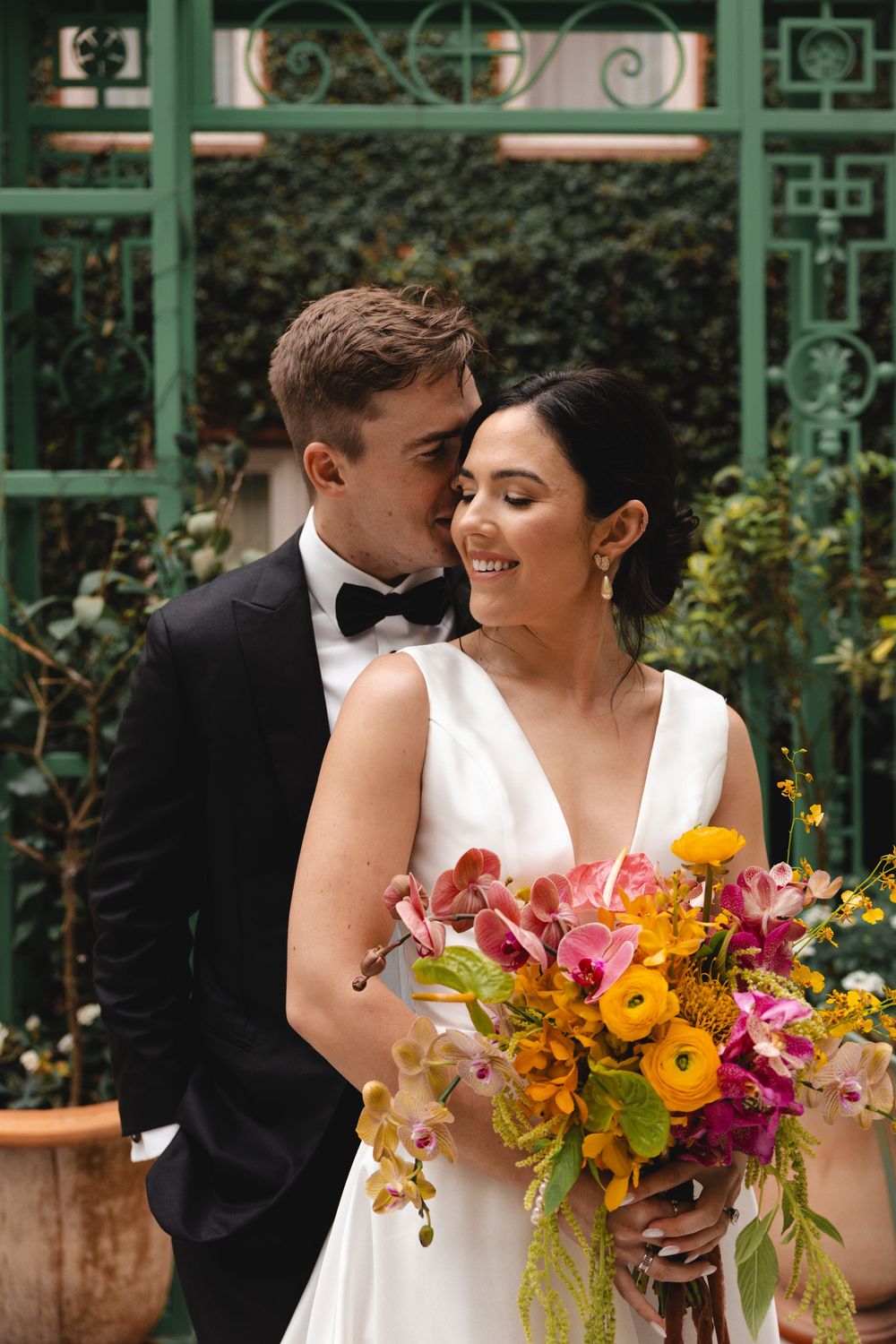 Bride holding colorful bouquet smiles as groom in black tuxedo kisses her cheek before green garden gate.