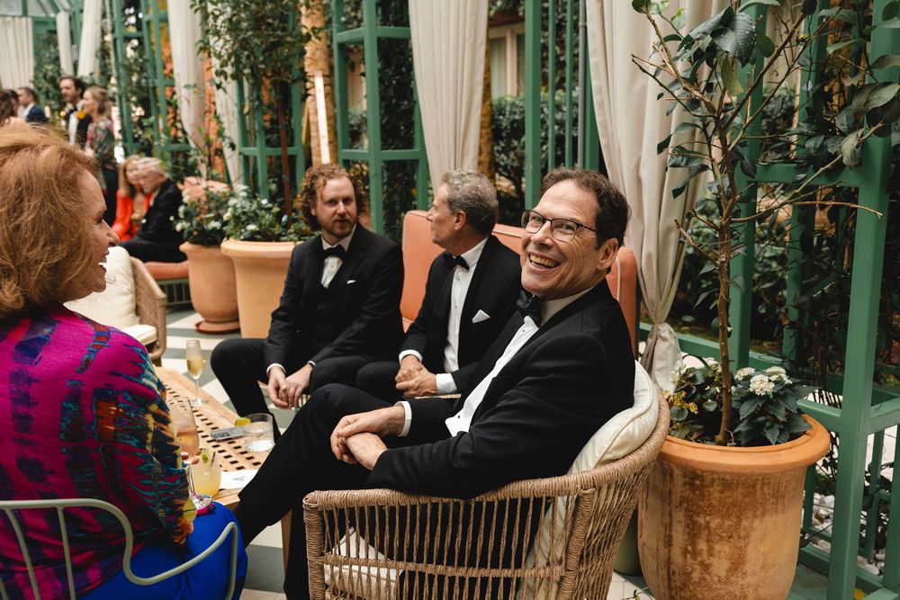 Men in tuxedos socializing at an elegant indoor garden party with lush greenery and warm ambient lighting.