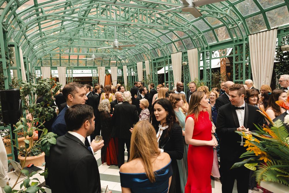 Elegant guests mingle at a glamorous greenhouse gala event, dressed in formal attire amid lush greenery.