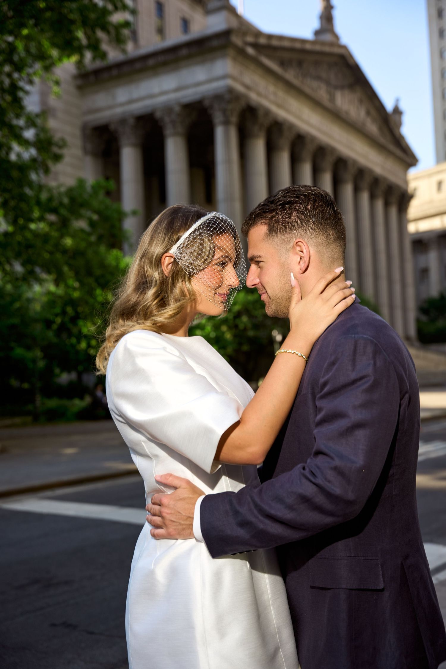 Bride in white dress and veil embraces groom in navy suit outside a neoclassical building on their wedding day.