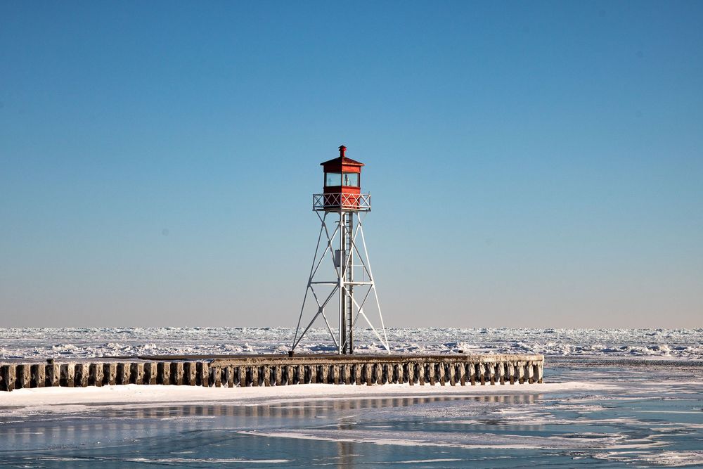 Red lighthouse on a snowy pier surrounded by frozen lake waters under a clear blue winter sky.