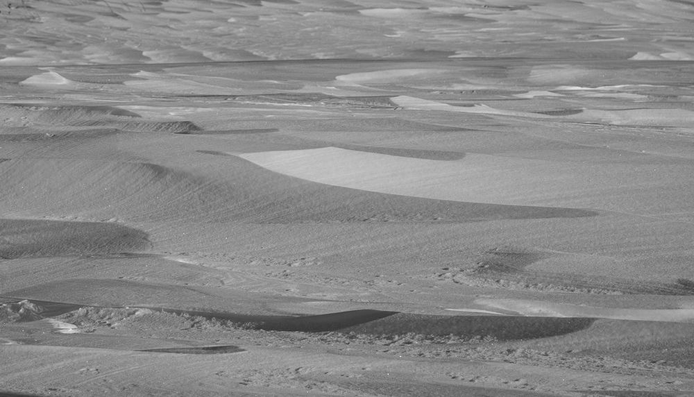 Black and white aerial view of vast desert sand dunes with rippling patterns and shadows across the landscape.
