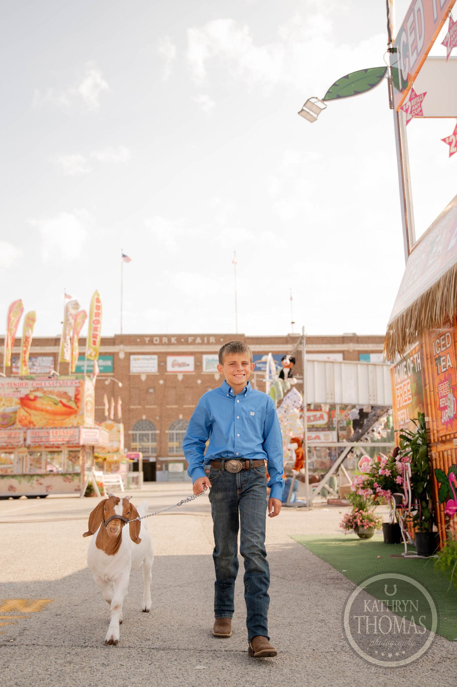 Young boy in blue shirt and jeans walks a goat at a county fair on a sunny day.