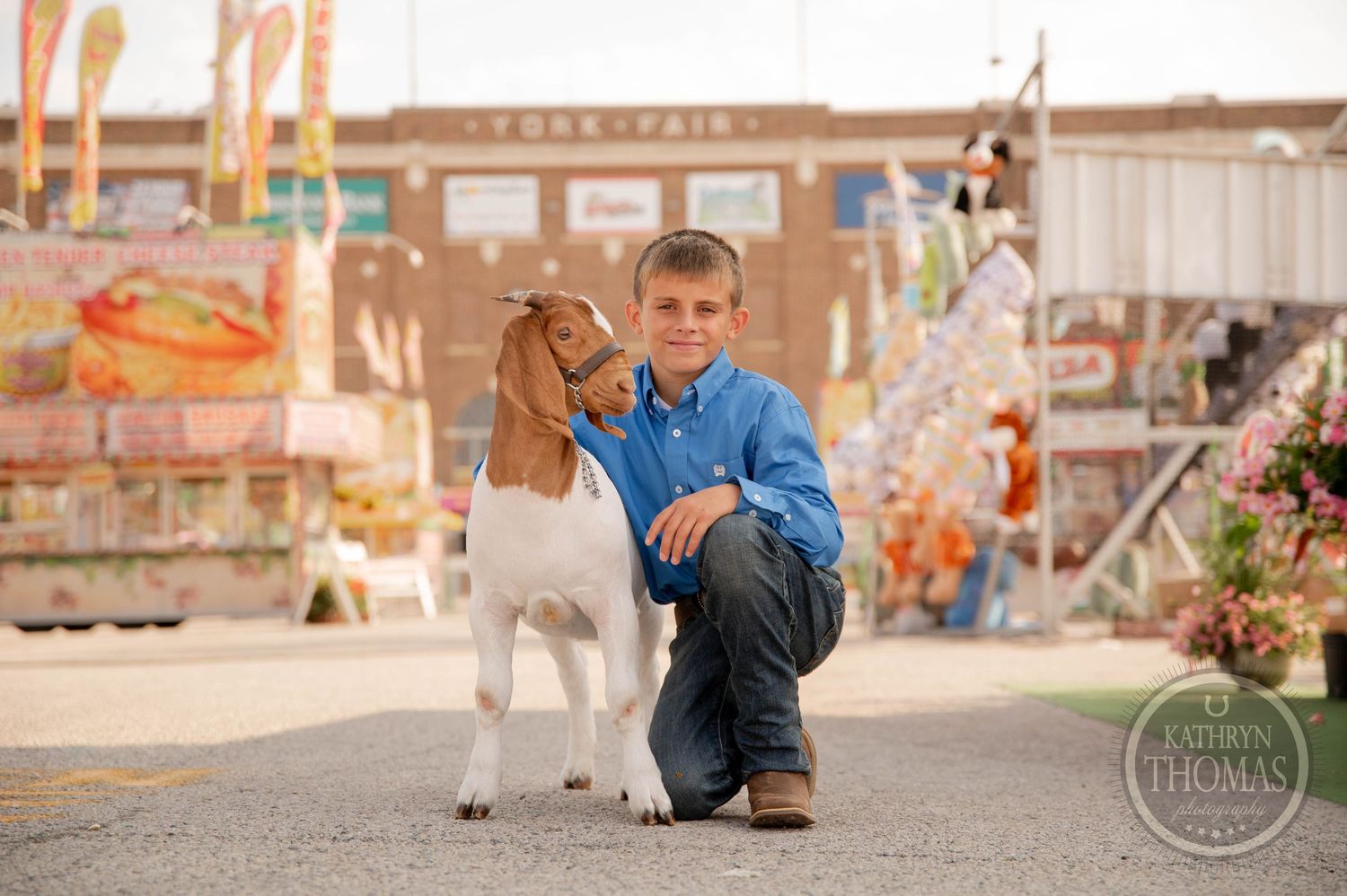 A boy in a blue shirt leads a goat at a county fair with colorful booths and a brick building in the background.