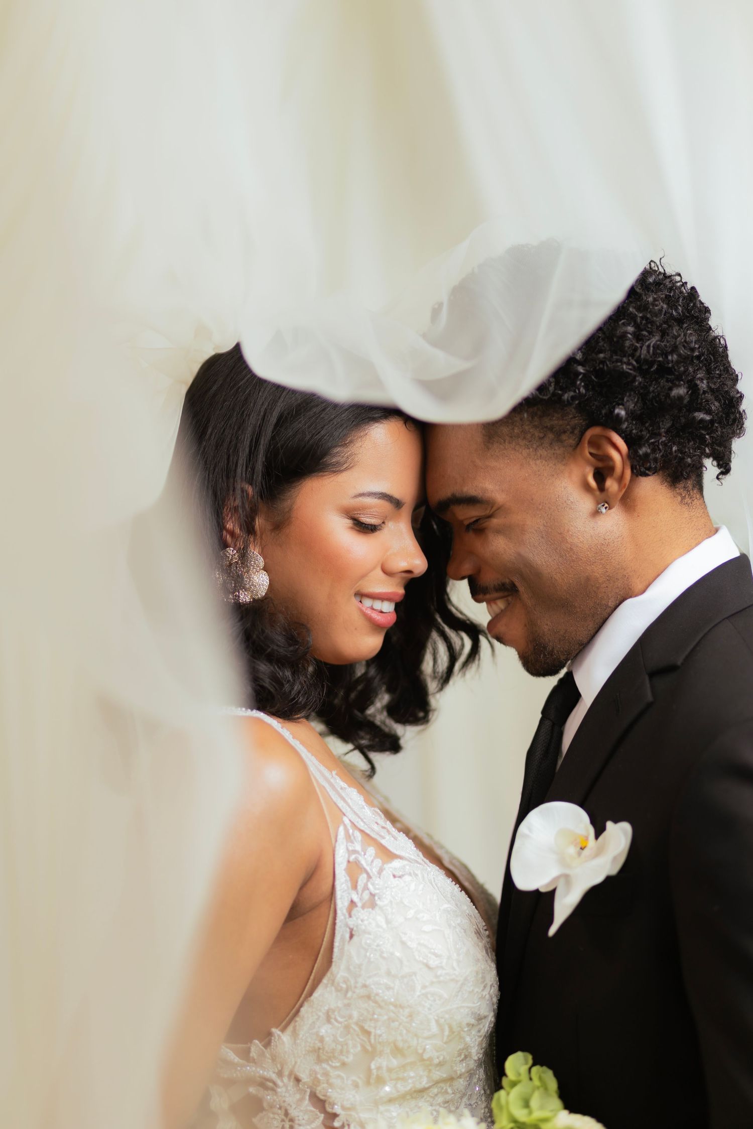 A joyful bride and groom share an intimate forehead touch under a flowing white veil on their wedding day.