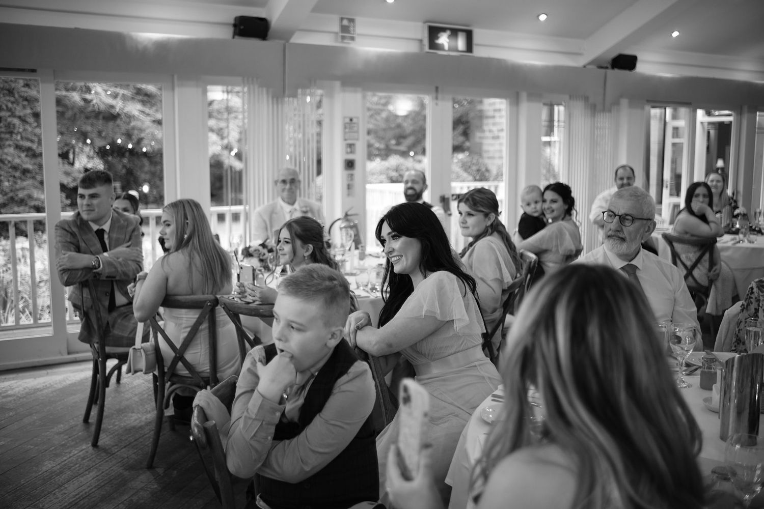 Black and white photo of wedding guests seated at tables in an elegant venue with large windows.