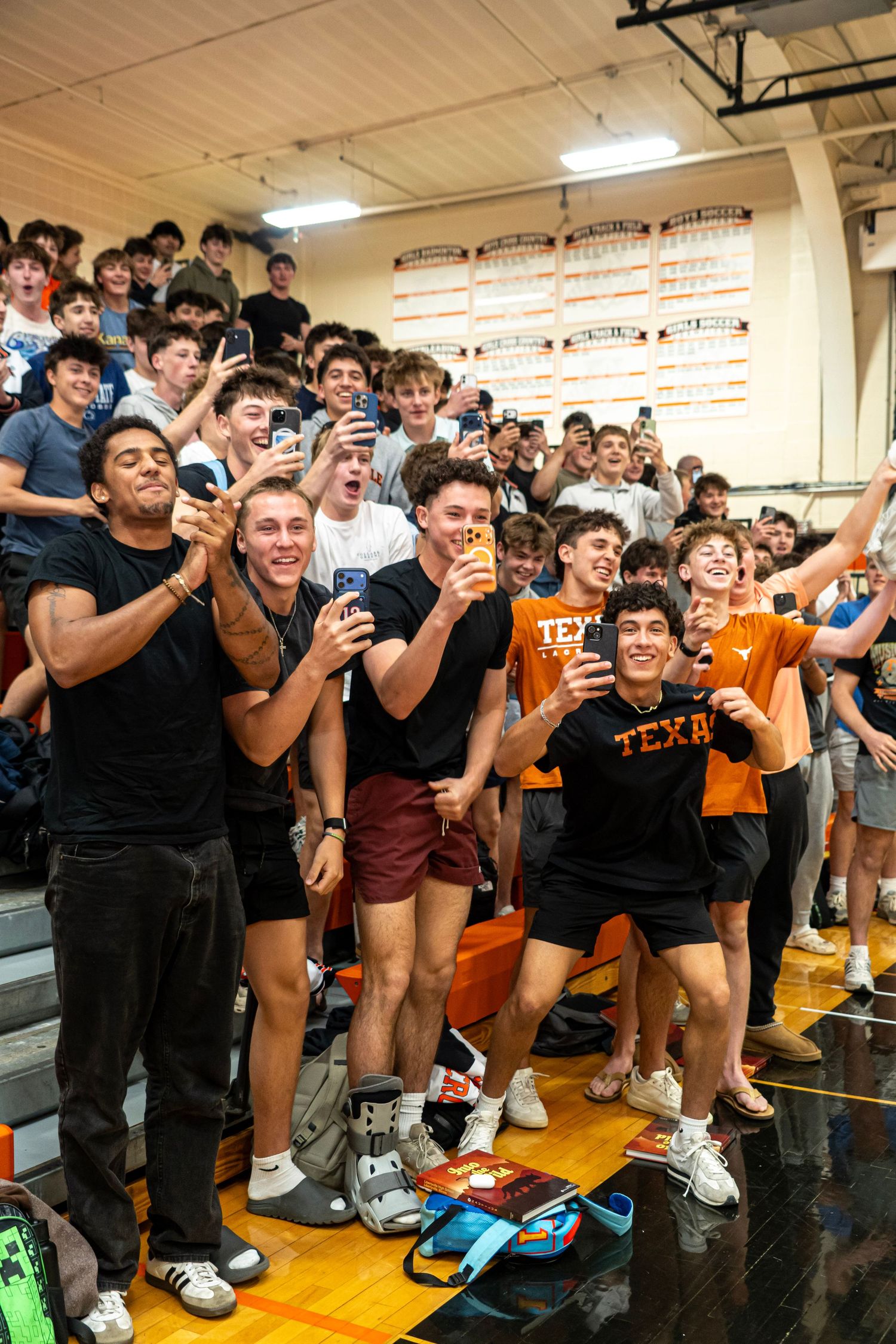 Excited crowd of young men in Texas Longhorns shirts cheering and taking selfies in a school gymnasium.