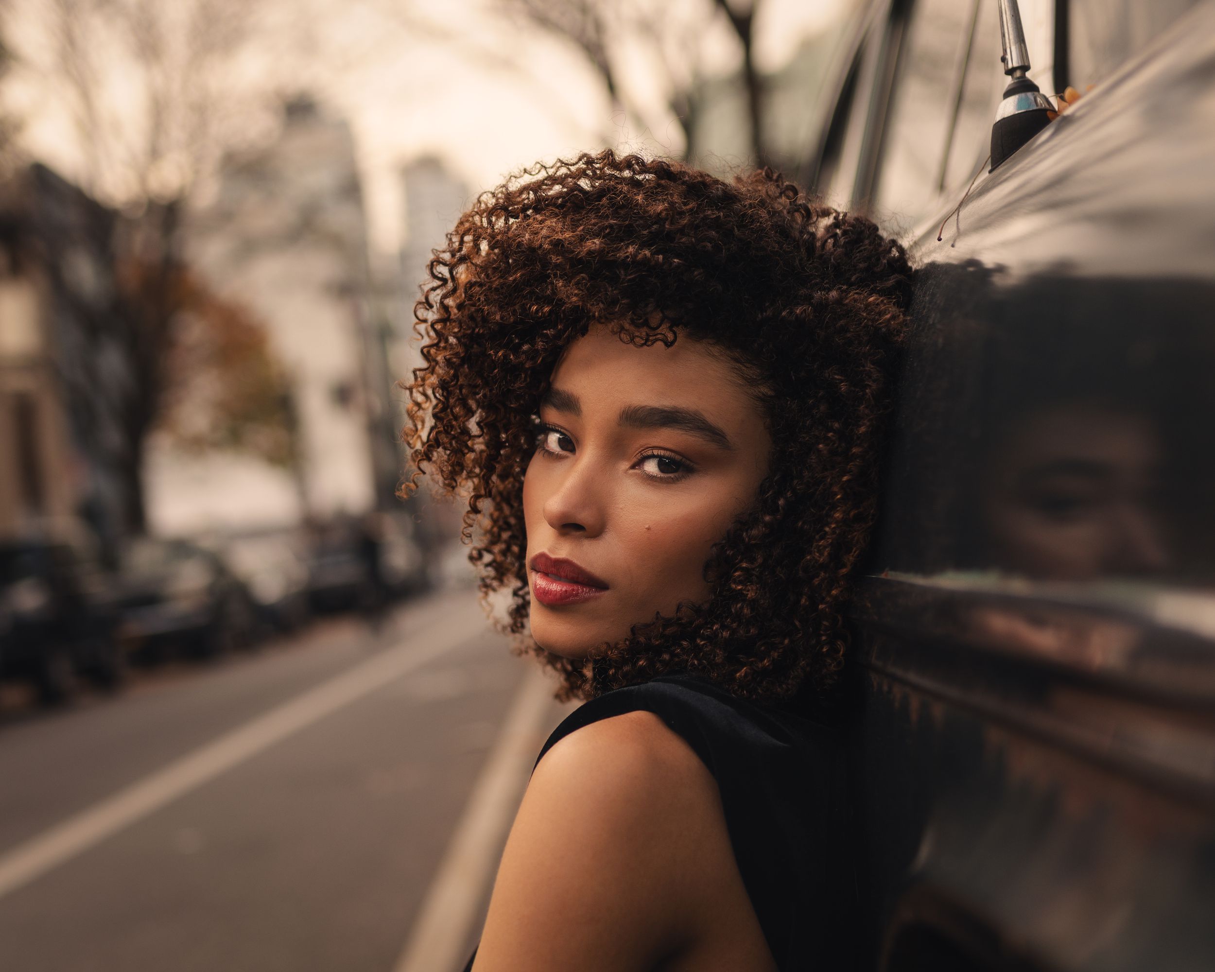 Young woman with curly auburn hair leaning against a dark vehicle on a city street, looking over her shoulder.