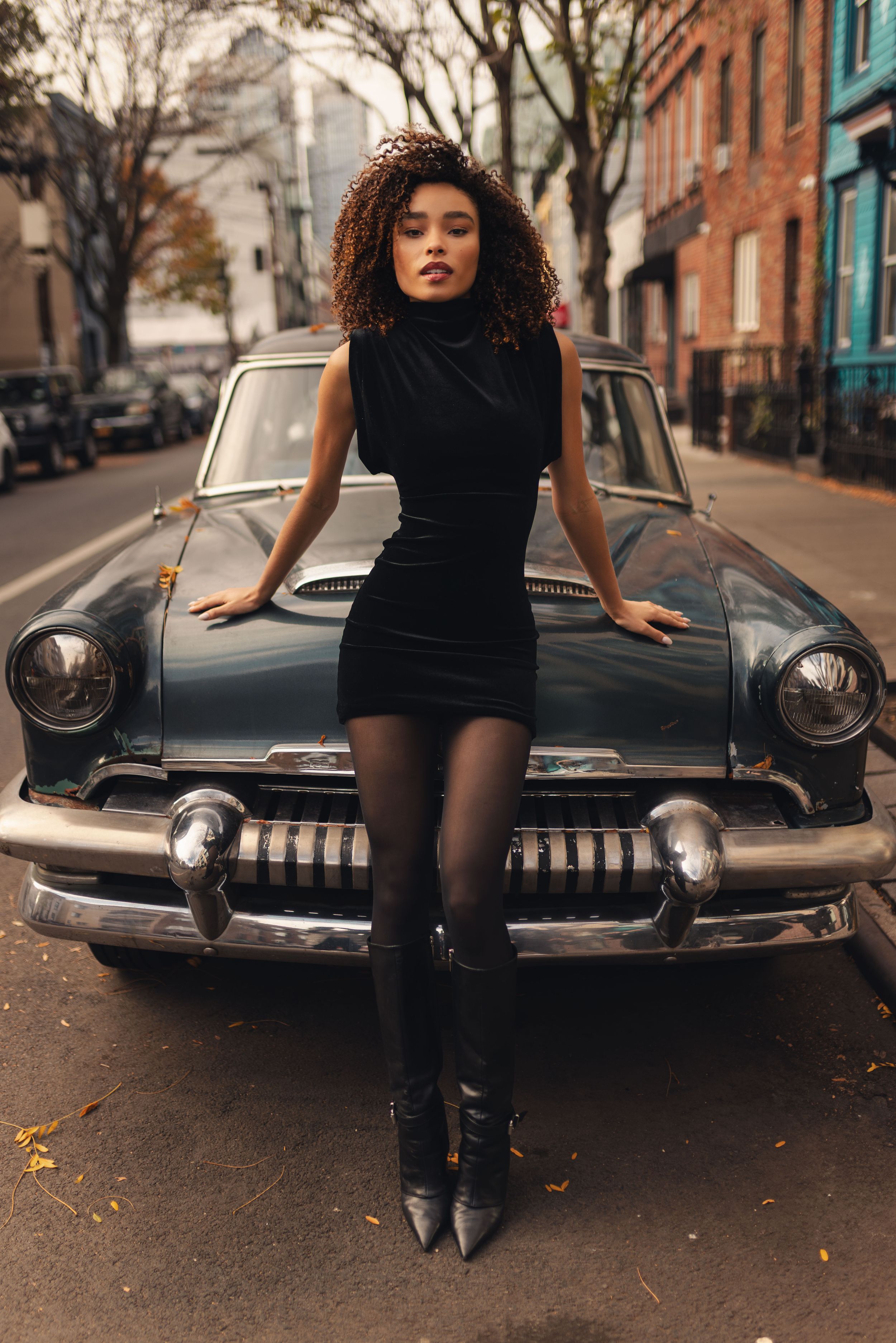 Young woman in black dress and tights posing on vintage classic car on urban street.