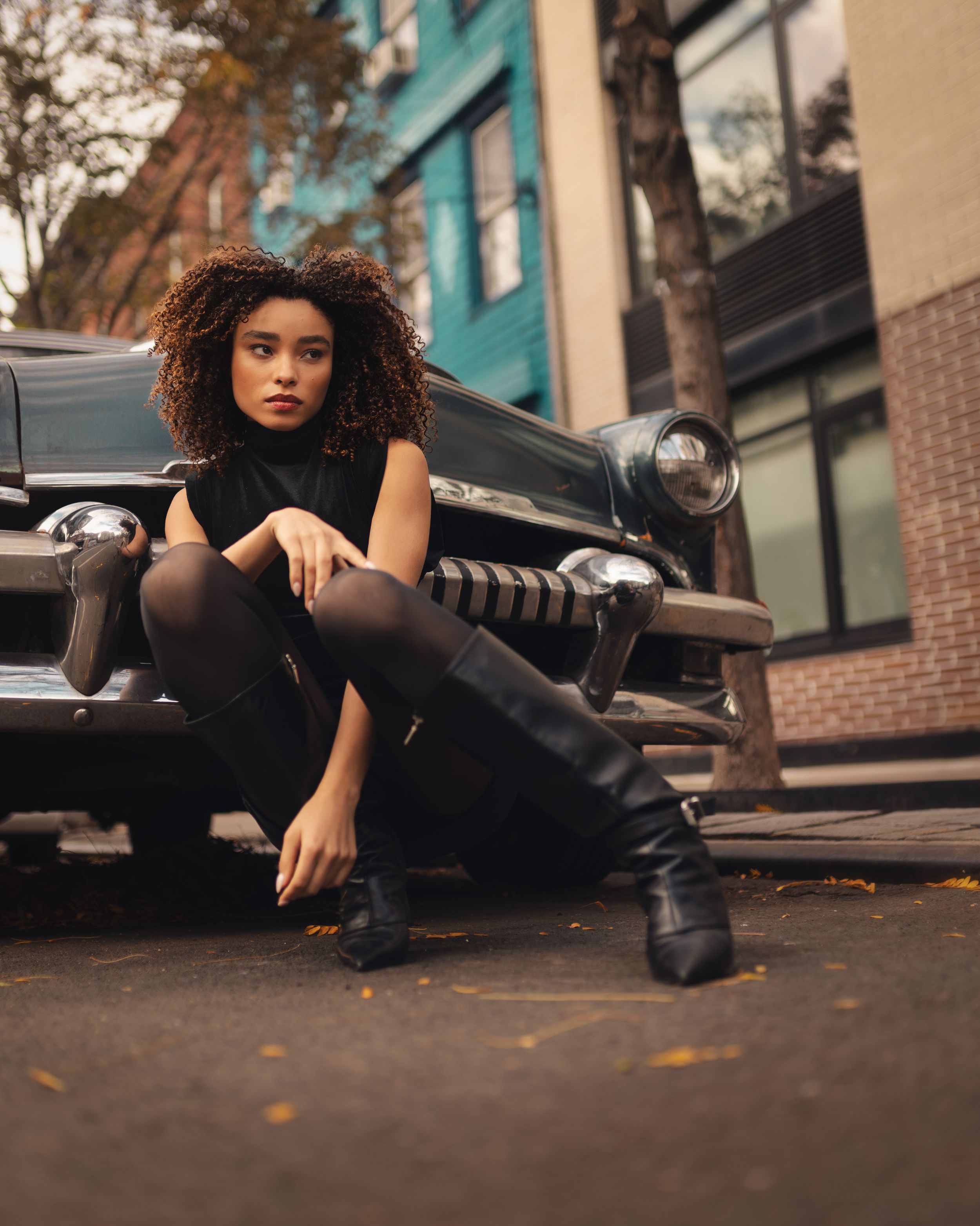 A young woman with curly hair poses in all-black outfit and knee-high boots beside a vintage car on an urban street.