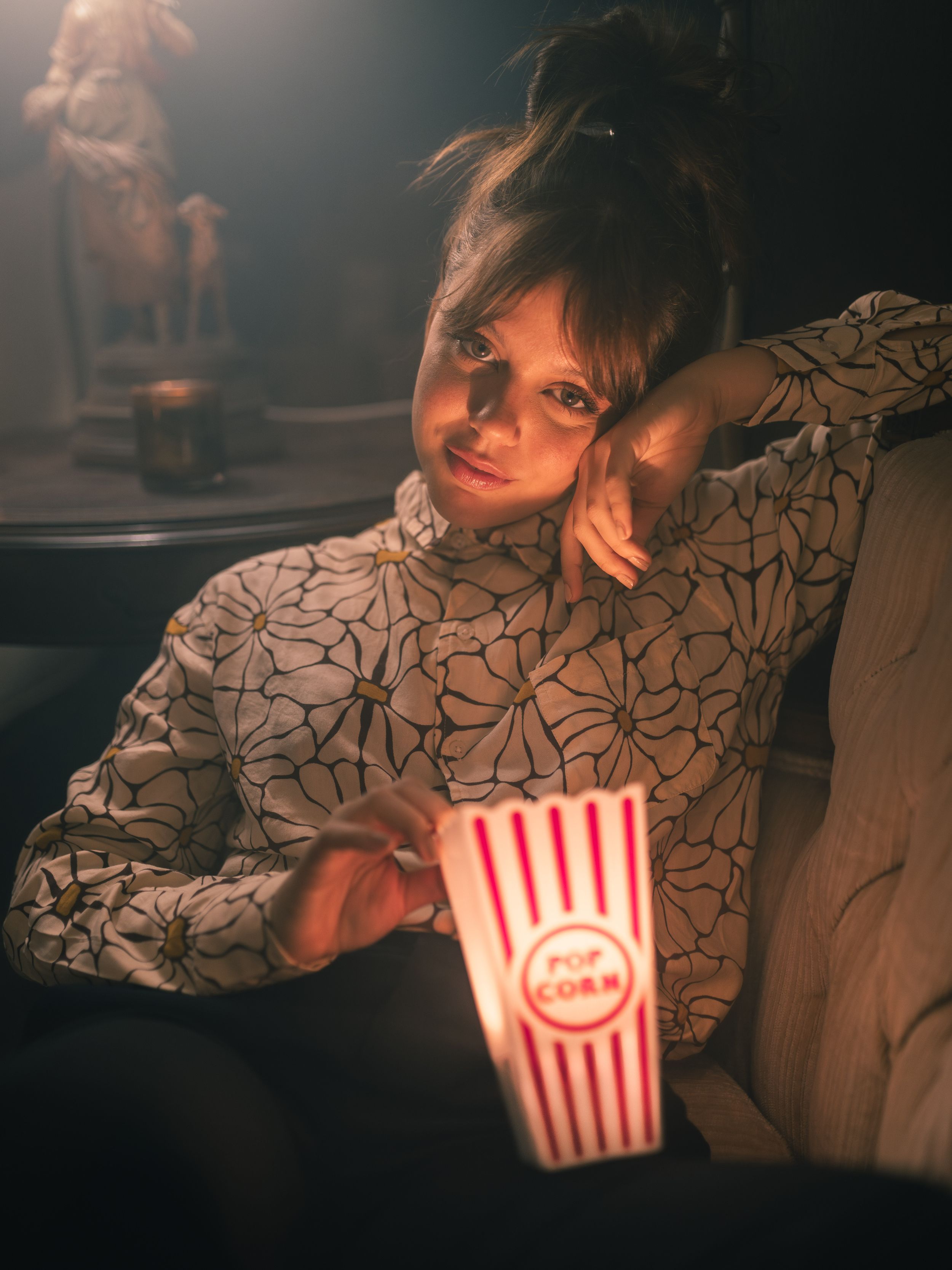 Woman relaxing in a dark cinema holding a striped popcorn bucket, wearing a floral blouse, lit by warm light.