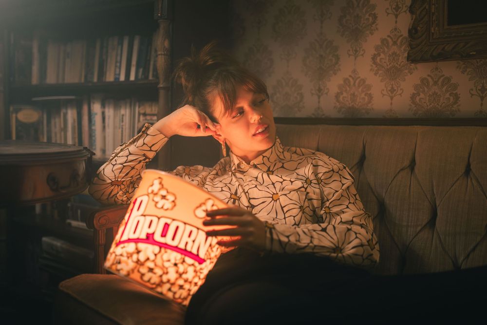 Young person relaxing on a couch watching a movie while holding a large popcorn bucket in a cozy dimly lit room.
