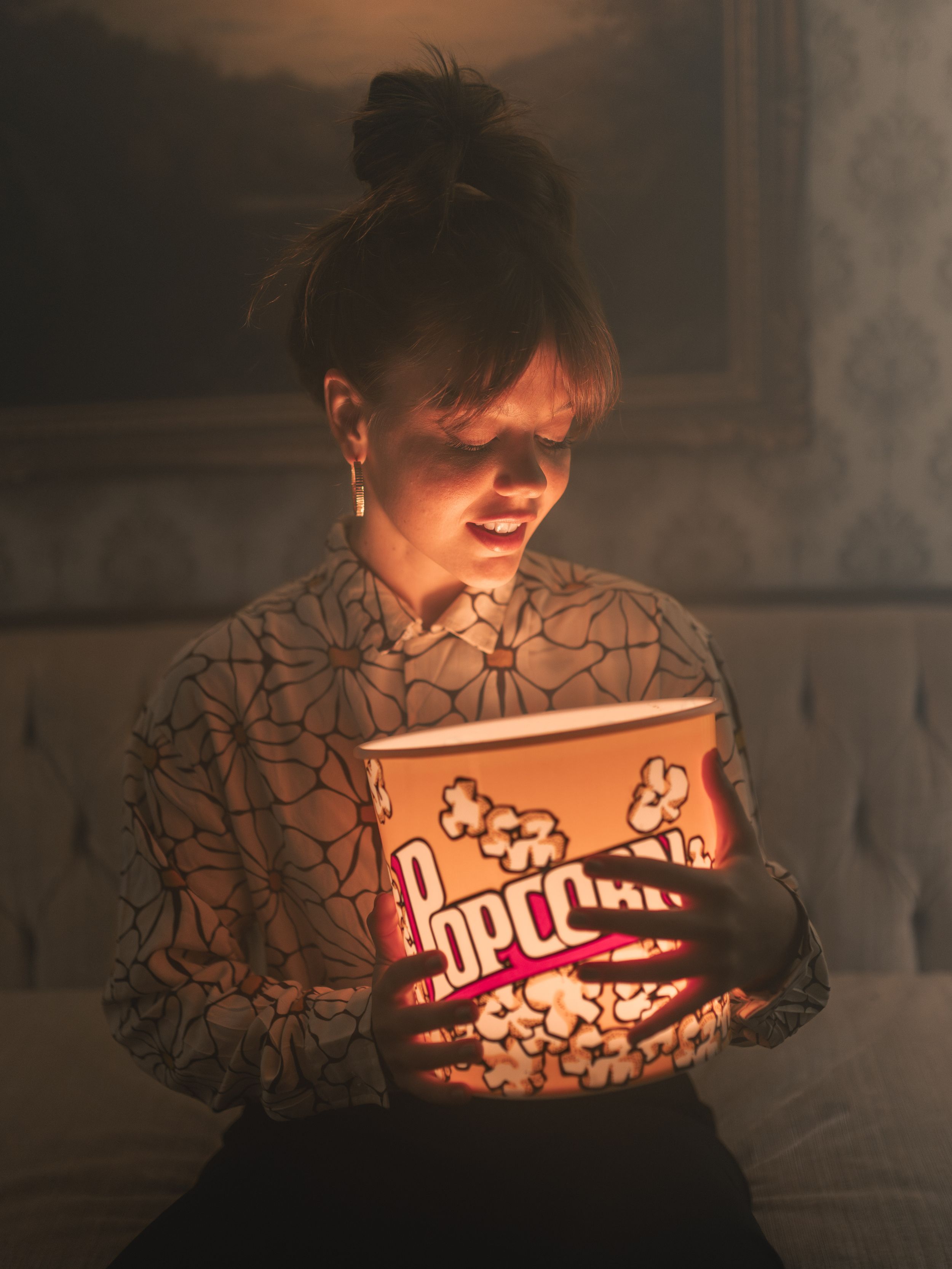 Woman holding glowing popcorn bucket in dark room wearing patterned blouse with hair in bun.