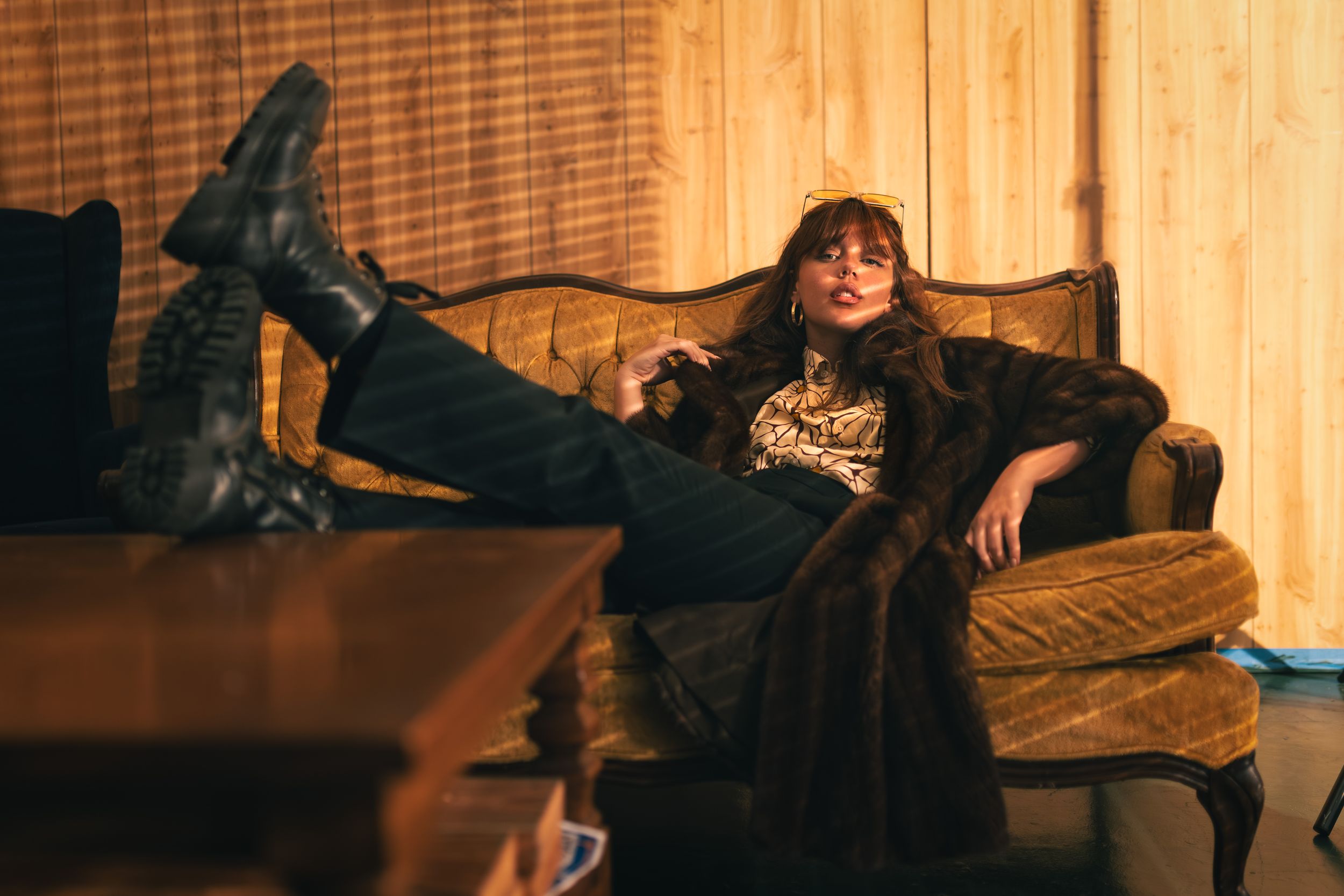 Woman in fur coat and boots lounging on vintage velvet chair in warm, moody retro-styled room.